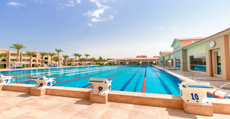 Aqua Vista Resort offers multiple outdoor pools, sun loungers, and umbrellas beneath clear blue skies—perfect for ein erfolgreiches Trainingslager in den Wintermonaten—with buildings and the sea in the background.