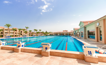 Aqua Vista Resort offers multiple outdoor pools, sun loungers, and umbrellas beneath clear blue skies—perfect for ein erfolgreiches Trainingslager in den Wintermonaten—with buildings and the sea in the background.