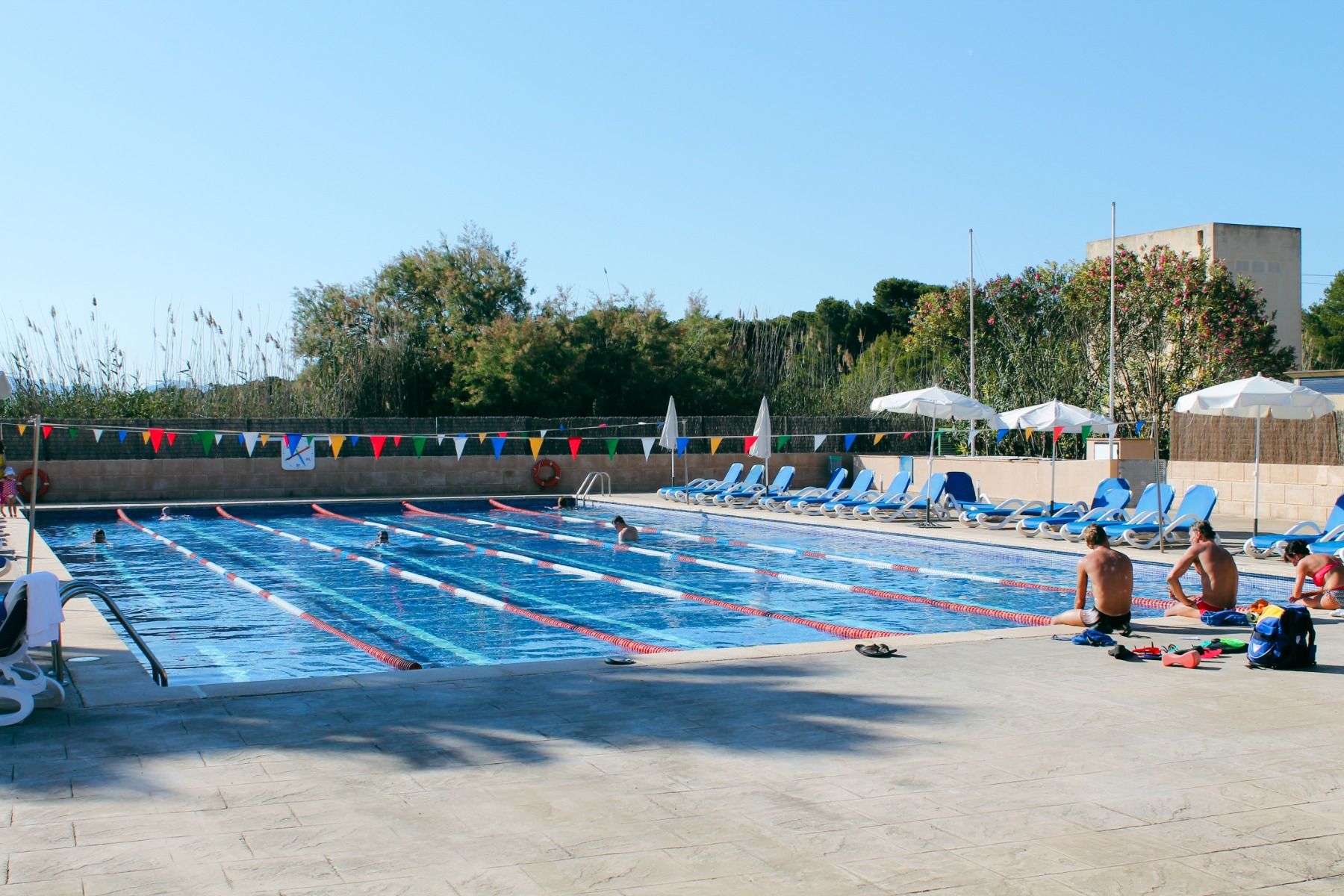 Schwimmbecken im Freien mit Schwimmern, blaue Liegen und Sonnenschirme am Beckenrand und bunten Wimpeln über dem Wasser.