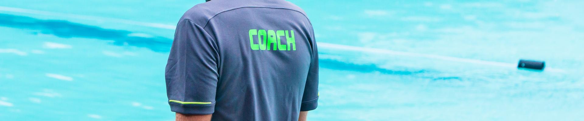 Rückansicht eines Trainers mit der Aufschrift "COACH" auf dem Shirt vor einem Schwimmbecken.