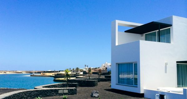 Moderne weiße Hotelvilla mit Panoramafenstern und überdachter Terrasse, umgeben von schwarzem Sand und Blick auf eine ruhige Lagune.