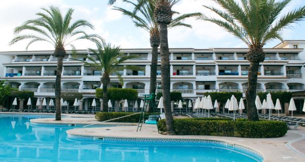Hotelgebäude im mediterranen Stil mit weißen Balkonen, Palmen und einem großen Poolbereich mit Sonnenliegen und Sonnenschirmen.