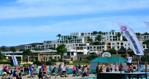 Outdoor-Fitnesskurs mit einer Gruppe von Menschen, die gemeinsam auf Steppern trainieren, vor der Kulisse einer weißen, terrassenförmigen Hotelanlage.