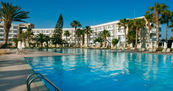 Große Poollandschaft umgeben von Palmen und Sonnenliegen vor dem Louis Phaethon Beach Hotel mit der weißen Hotelanlage im Hintergrund.