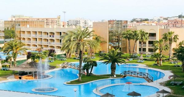 Weitläufiger Hotelgarten mit geschwungenem Pool, Palmen und einer Brücke, umgeben von Hotelgebäuden.