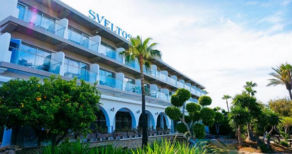 Außenansicht des Sveltos Hotel mit modernen Balkonen, Palmen und gepflegtem Gartenbereich unter blauem Himmel.
