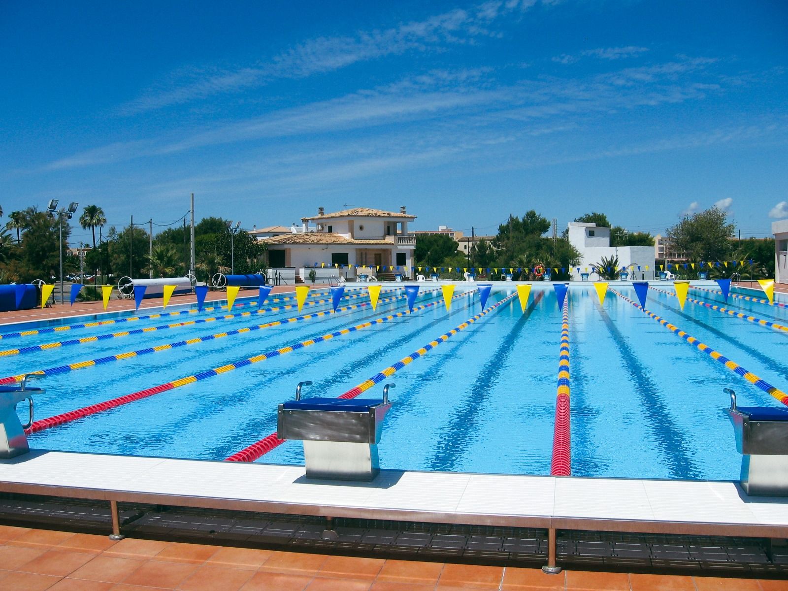 Ein großes, einladendes Freibad mit klaren blauen Bahnen, umgeben von Sonnenschirmen und einer sonnigen, mediterranen Umgebung mit Palmen im Hintergrund.