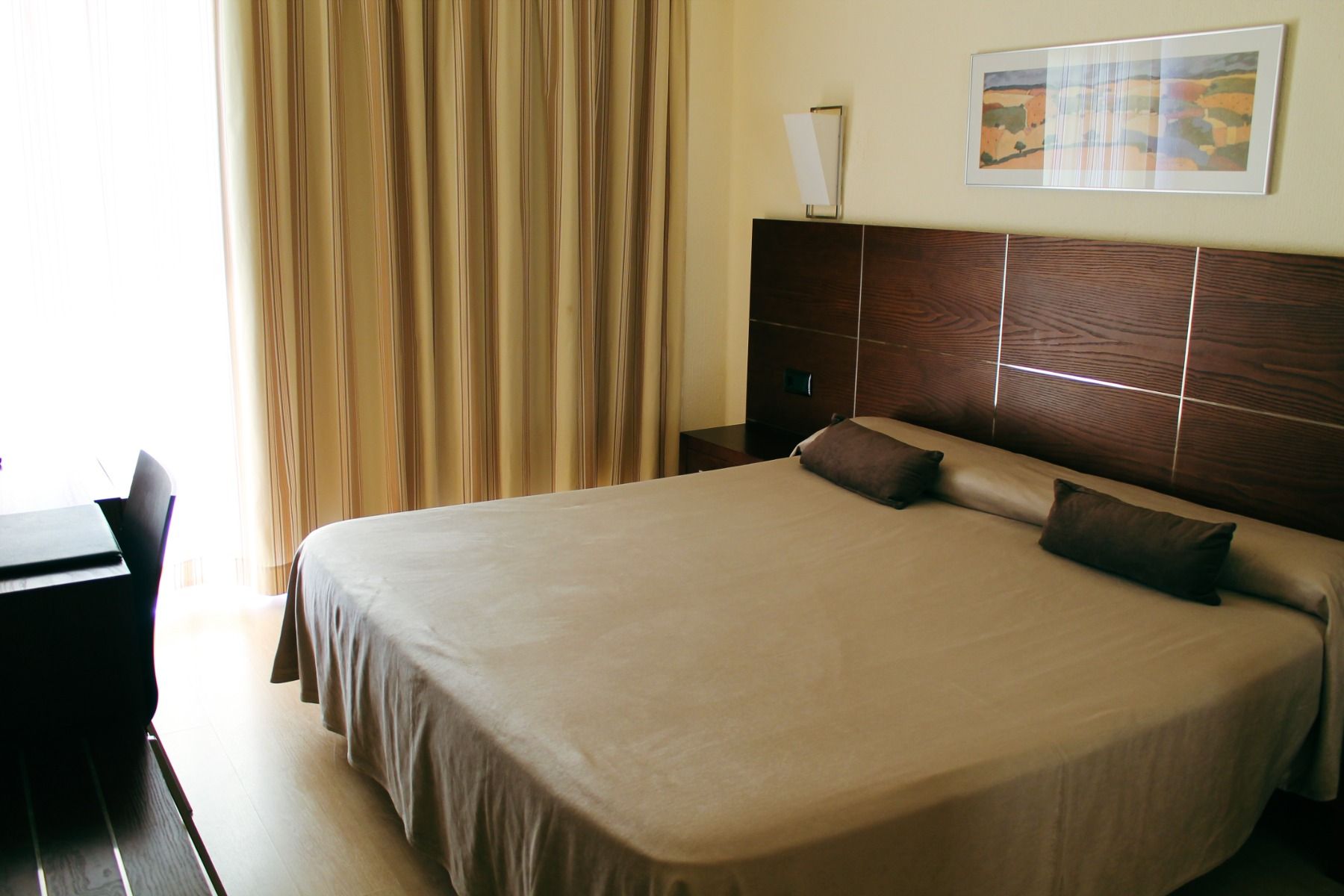 Ein gemütlich eingerichtetes Hotelzimmer mit einem Doppelbett, dunkelbraunem Holzrahmen und passenden Kopfkissen, sowie lichtdurchfluteten Vorhängen, die einen warmen Ton erzeugen.