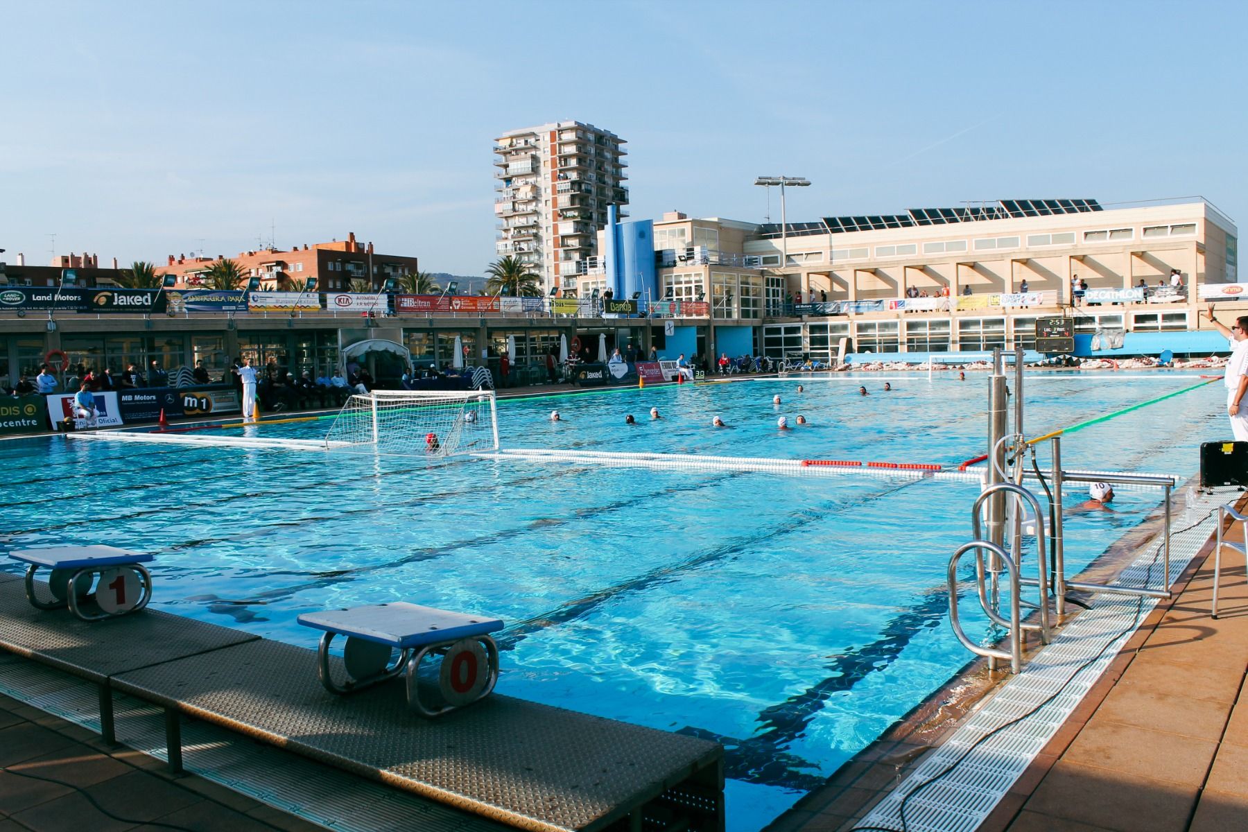 Ein großes Freibad mit Wasserballtoren, in dem Sportler trainieren. Im Hintergrund sind Zuschauertribünen und ein mehrstöckiges Gebäude zu sehen.