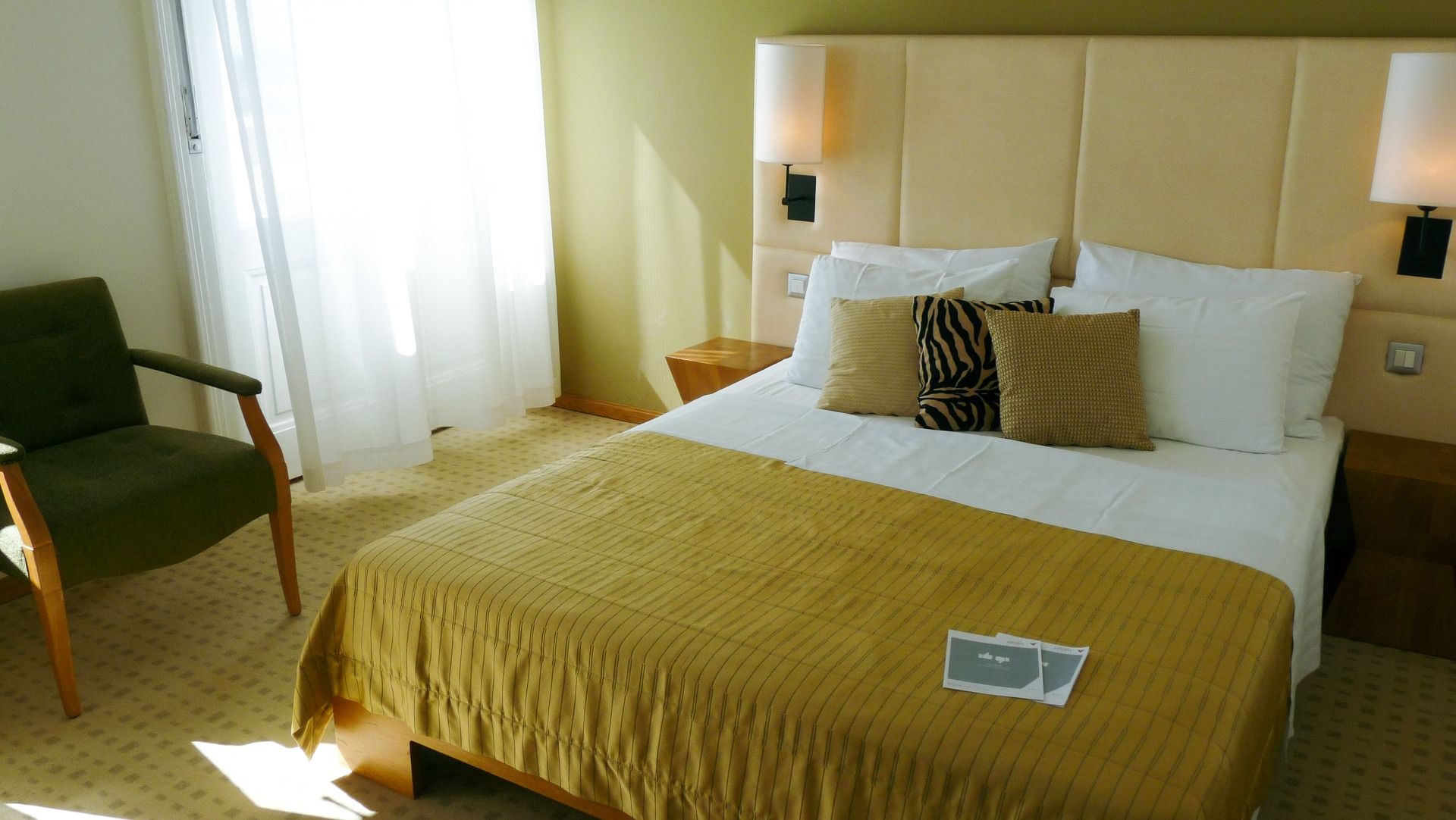 Modernes Doppelbett mit goldfarbener Bettdecke und dekorativen Kissen in einem sonnendurchfluteten Zimmer im Hotel Astoria