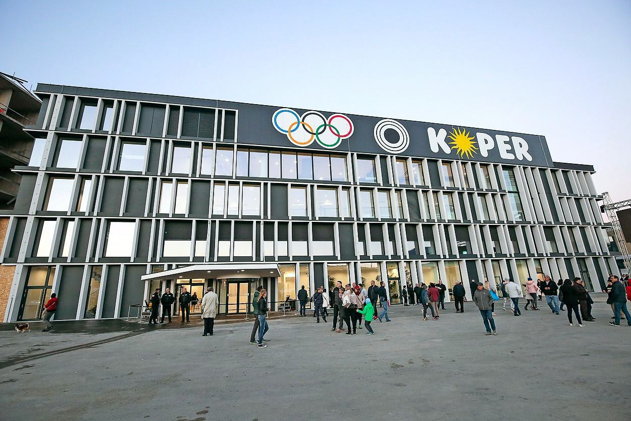 Moderne Gebäudefassade mit olympischen Ringen und dem Schriftzug Koper, umgeben von Menschen.