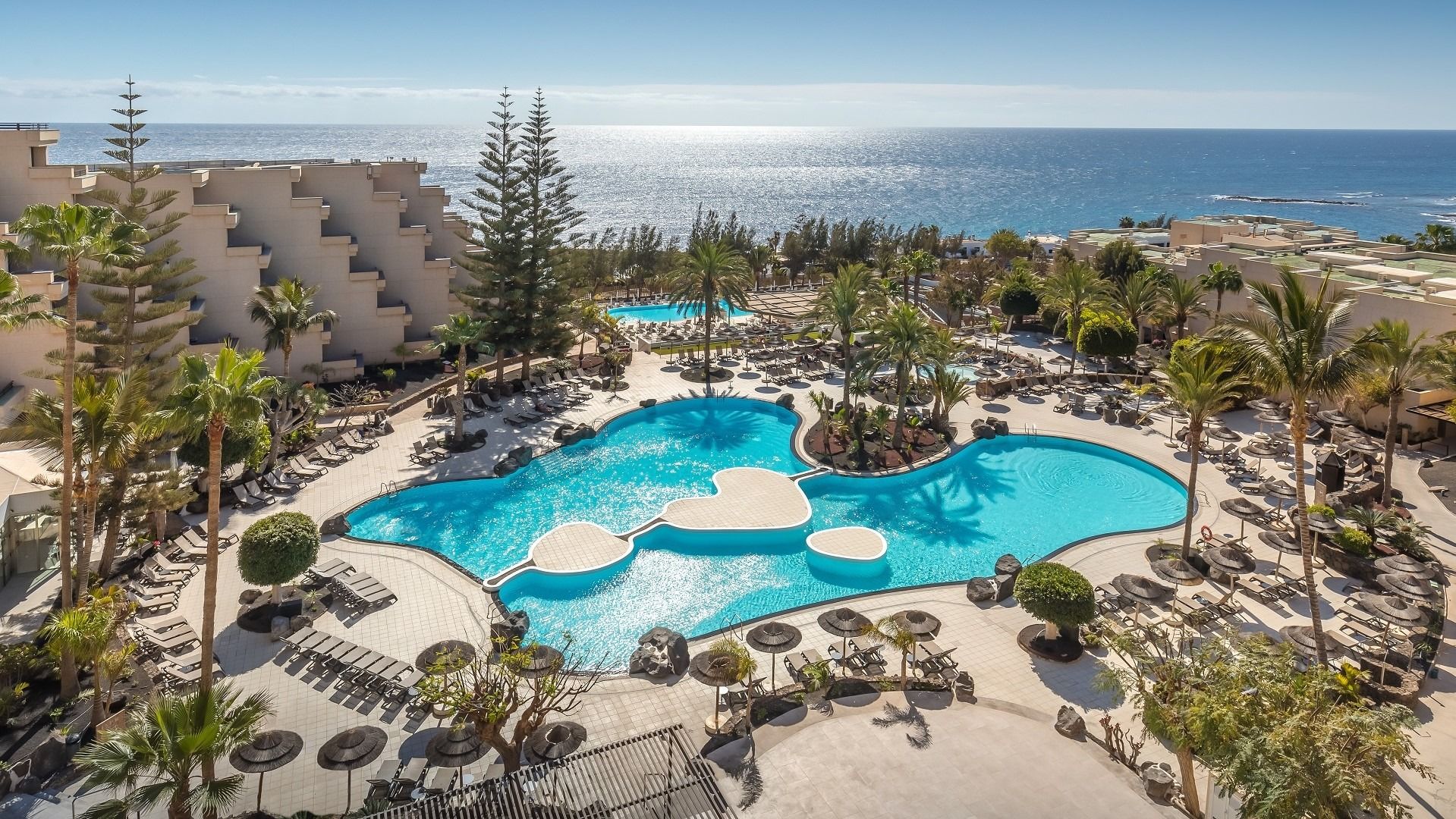 Luftaufnahme einer großen Poolanlage mit geschwungenen Formen, Palmen und Blick auf das Meer.