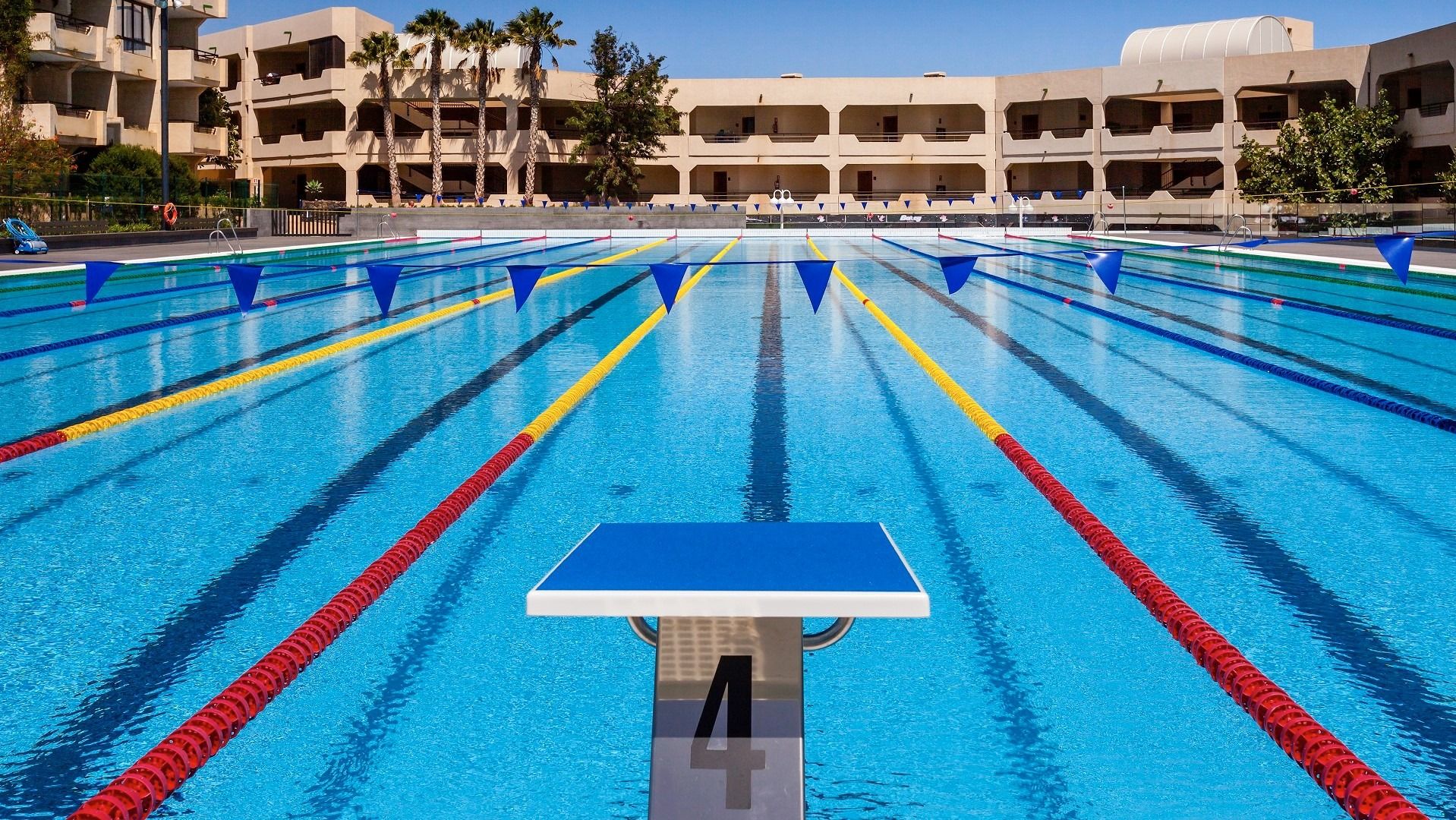 Nahaufnahme eines Startblocks an einem Schwimmbecken mit bunten Bahnmarkierungen und Hotelfassaden im Hintergrund.