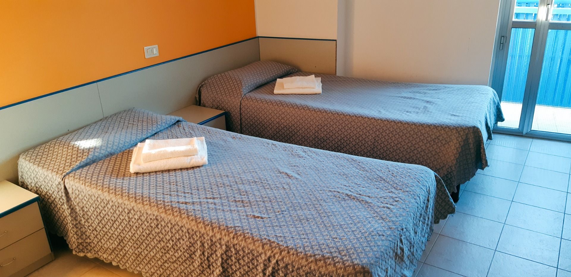 Zweibettzimmer mit schlichten grauen Überwürfen, orangefarbener Akzentwand und Zugang zu einem Balkon