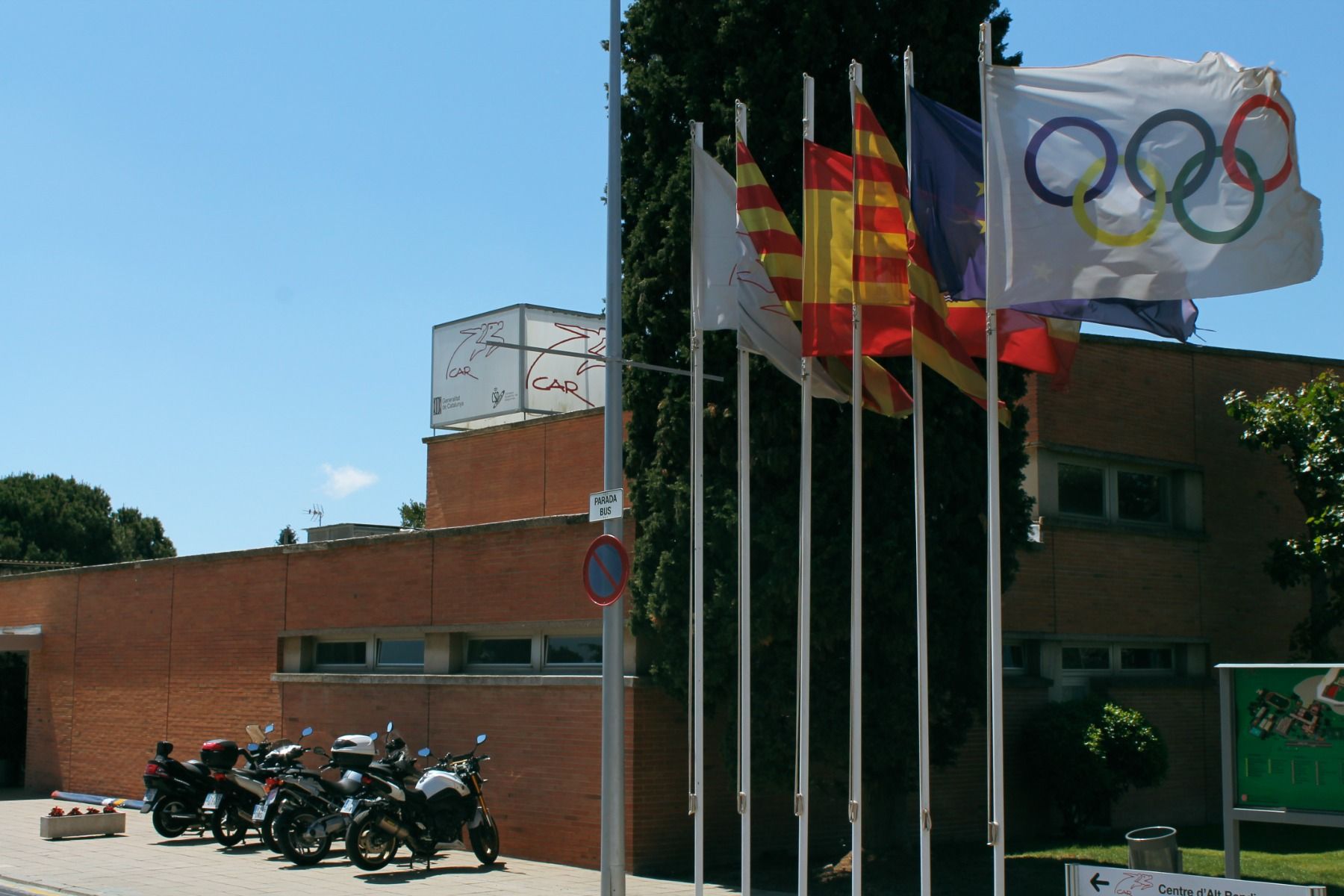 Eingang des CAR Centre d'Alt Rendiment mit wehenden Fahnen der Olympischen Spiele und regionalen Symbolen, daneben geparkte Motorräder.