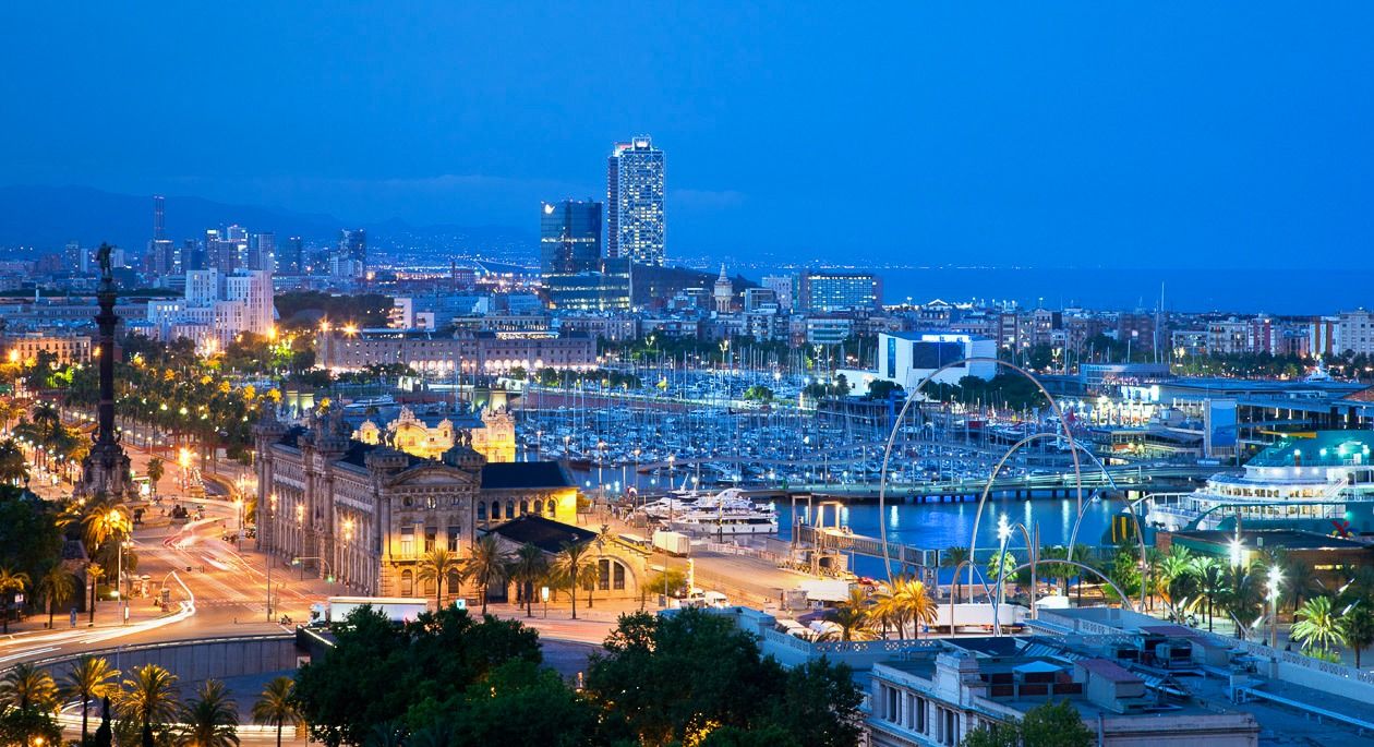 Nachtaufnahme der Stadt Barcelona, mit Blick auf den Hafen, beleuchtete Straßen und markante Hochhäuser im Hintergrund.