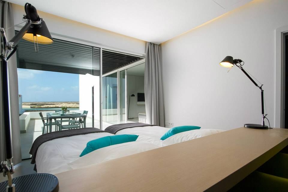 Helles Hotelzimmer mit zwei Einzelbetten, modernen Leselampen und einem Schreibtisch, mit direktem Zugang zu einer Terrasse mit Meerblick.