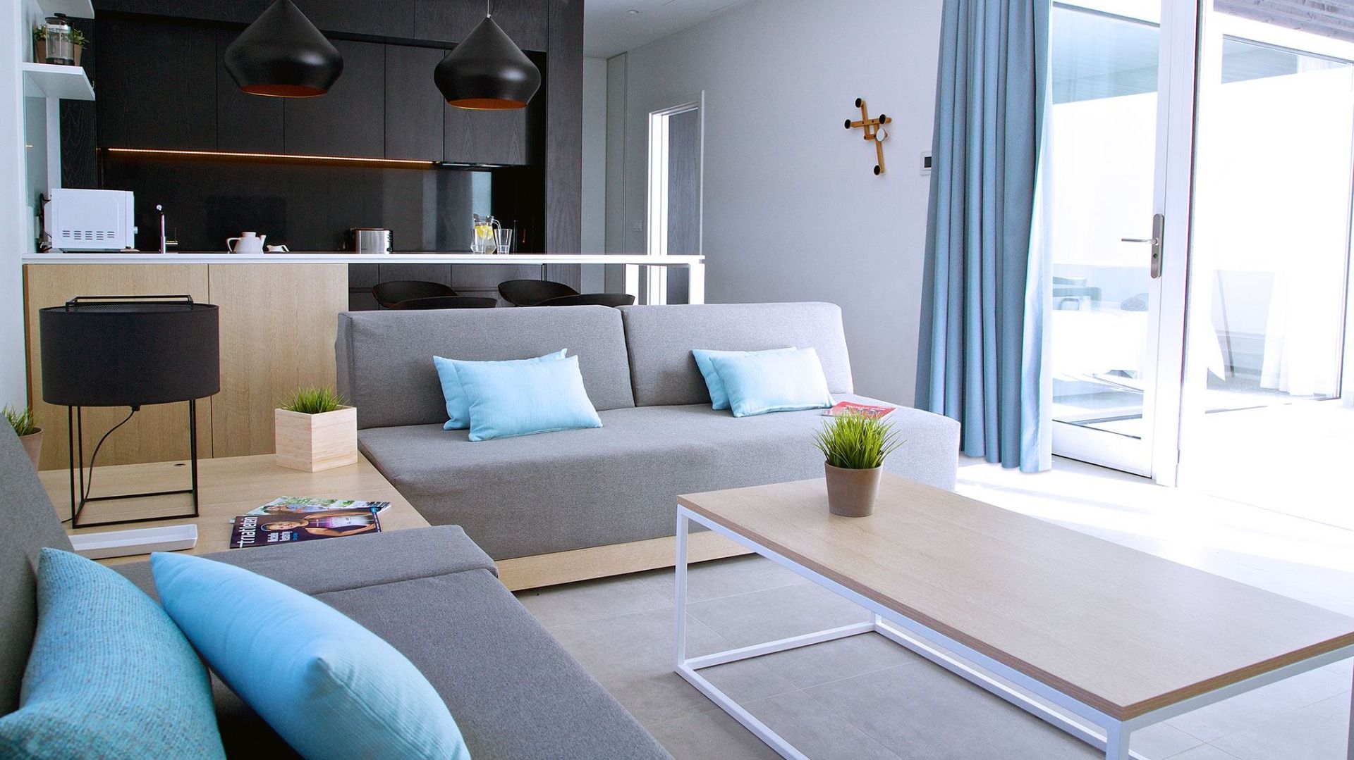 Stilvoll eingerichtetes Wohnzimmer mit grauen Sofas, blauen Kissen und einer offenen Küche mit schwarzen Hängelampen, moderner Dekoration und großen Fenstern zur Terrasse.