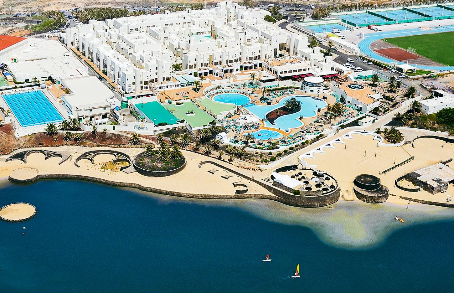 Luftaufnahme des Club La Santa Resorts mit mehreren Pools, Sportanlagen, einer blauen Lagune und weißen Hotelgebäuden in einer weitläufigen Landschaft.