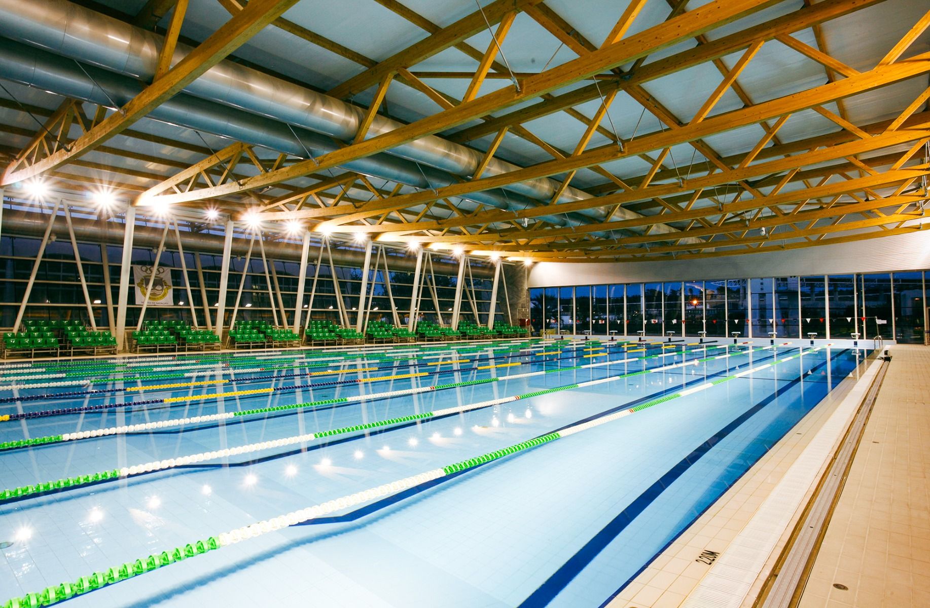 Beleuchtetes Hallenbad mit mehreren Bahnen und moderner Holzstruktur an der Decke.
