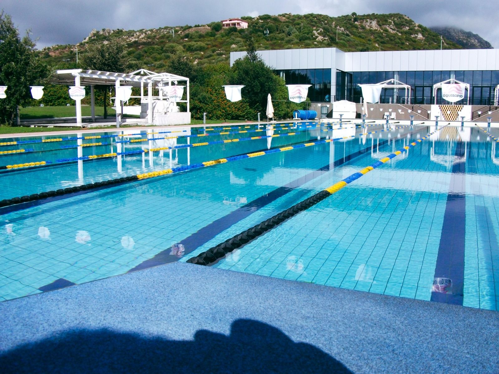 Außenpool mit klaren Schwimmbahnen, umgeben von grüner Landschaft, Pergolen und modernem Gebäude im Hintergrund