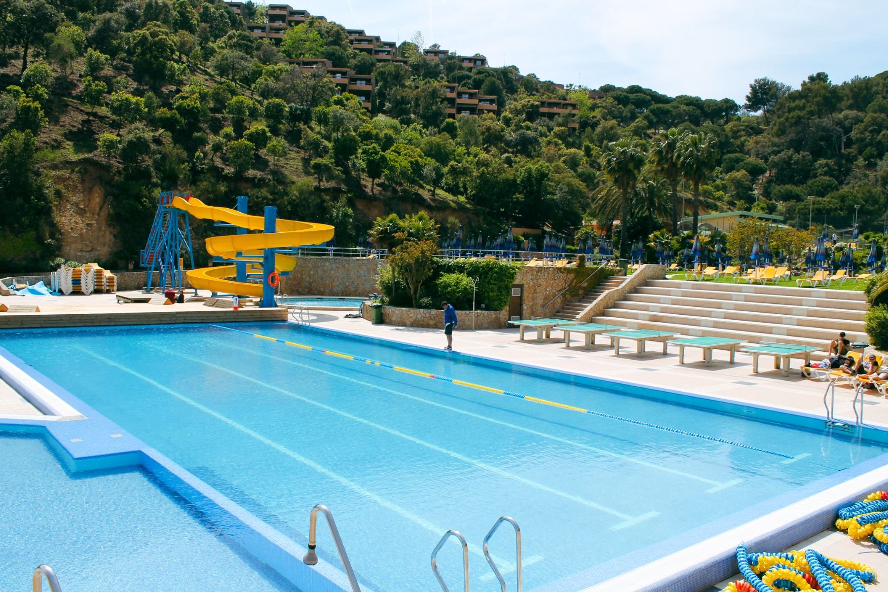 Outdoor-Pool mit Wasserrutsche, umgeben von grüner Landschaft und Sonnenliegen.