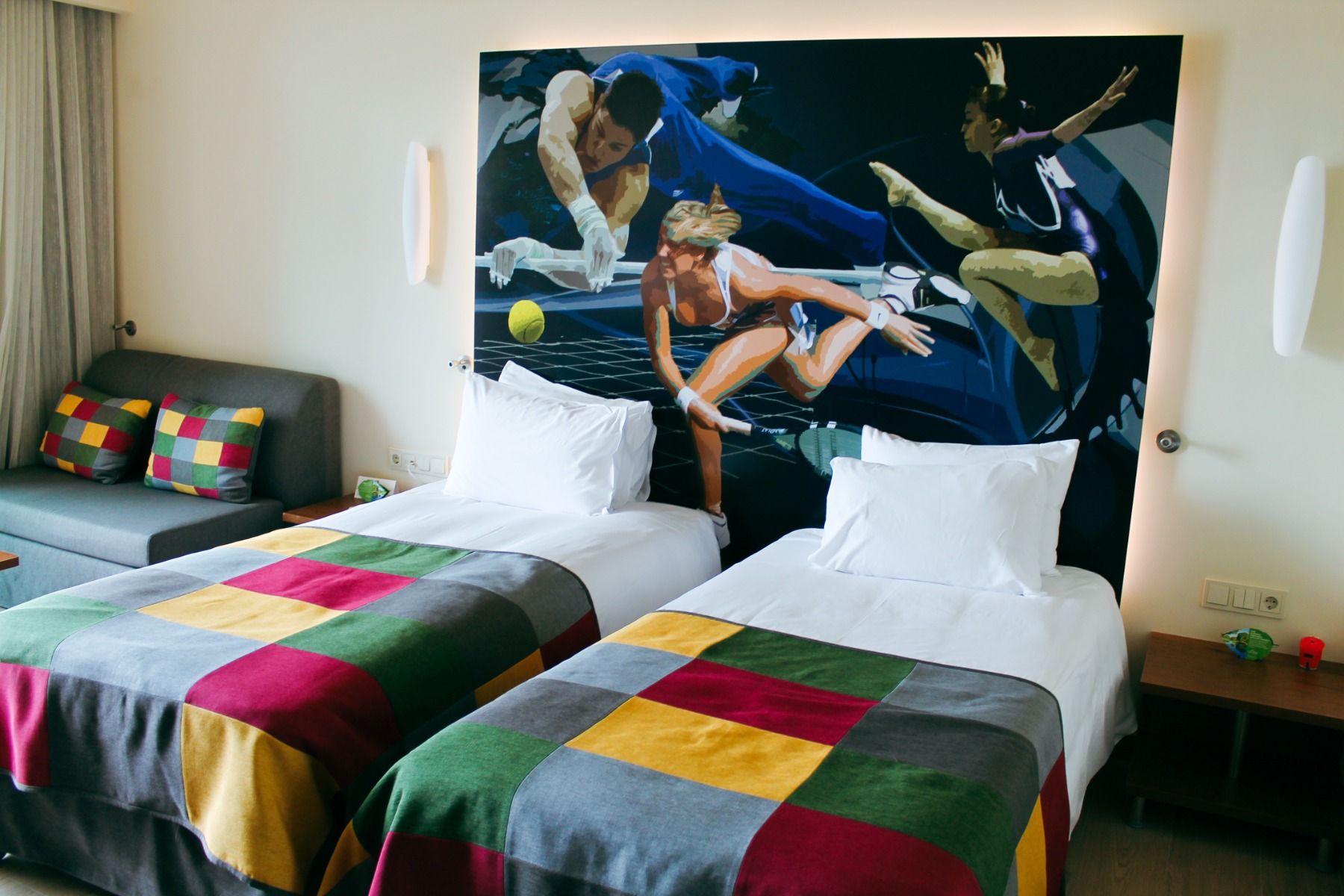 Stilvoll eingerichtetes Zimmer mit zwei Einzelbetten, bunter Tagesdecke und Wandkunst mit Sportmotiven.