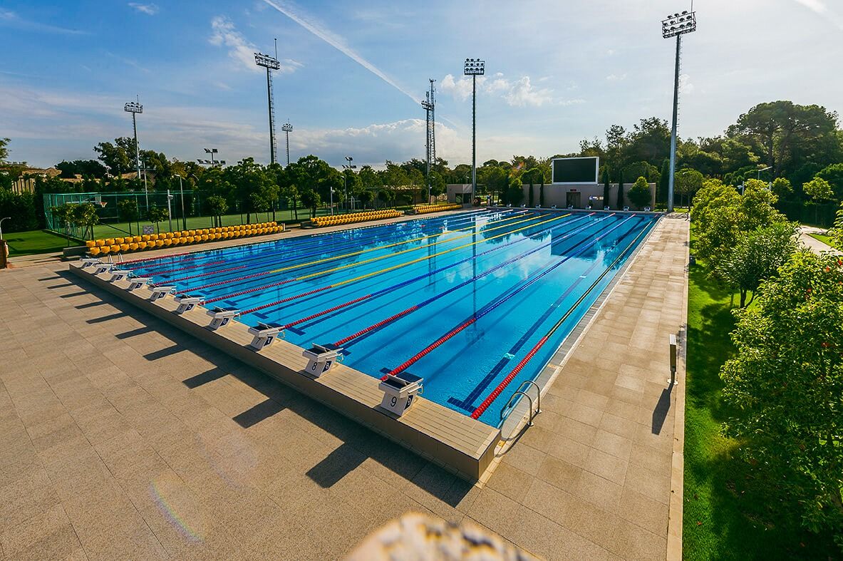 Freiluft-Sportbecken mit acht Schwimmbahnen und Startblöcken, umgeben von grünen Bäumen und Flutlichtmasten.