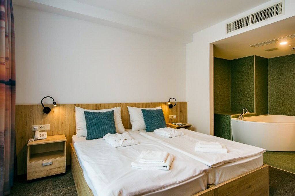 Stilvolles Hotelzimmer mit Doppelbett, zwei Nachttischen und einer modernen Whirlpool-Badewanne im Hintergrund. Komfortables und luxuriöses Ambiente.