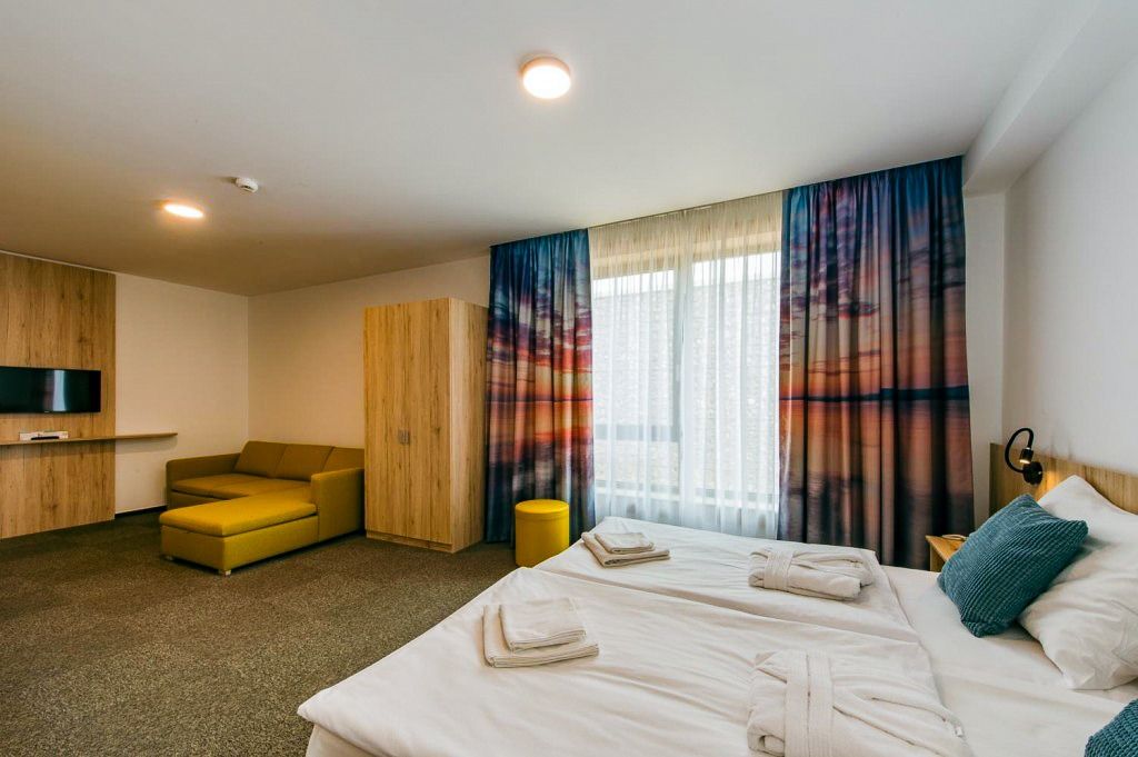 Großes, modernes Hotelzimmer mit einem Doppelbett, einer gelben Couch, und einer beeindruckenden Landschaft auf den Vorhängen. Minimalistisches und warmes Design.
