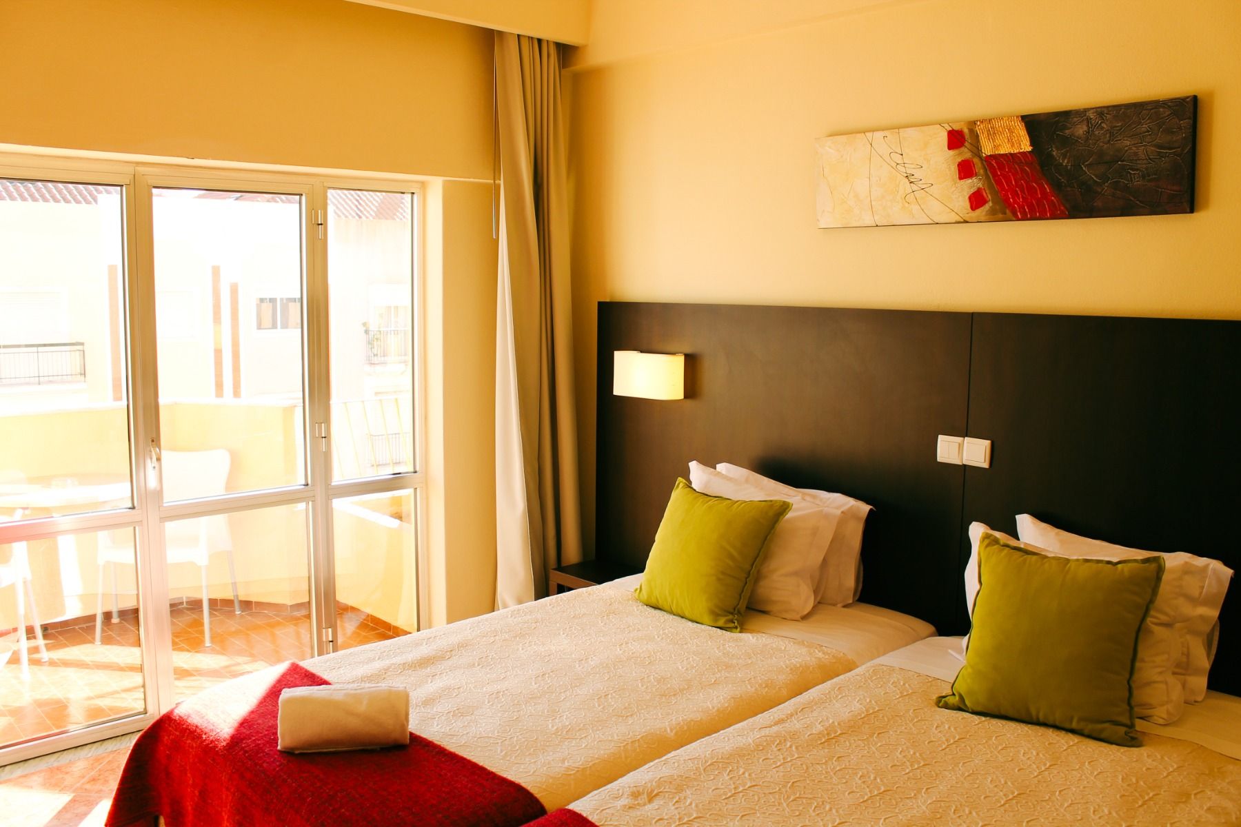 Hotelzimmer mit einem Doppelbett, grünen Kissen, roter Decke und Balkon mit Glasfenstern.