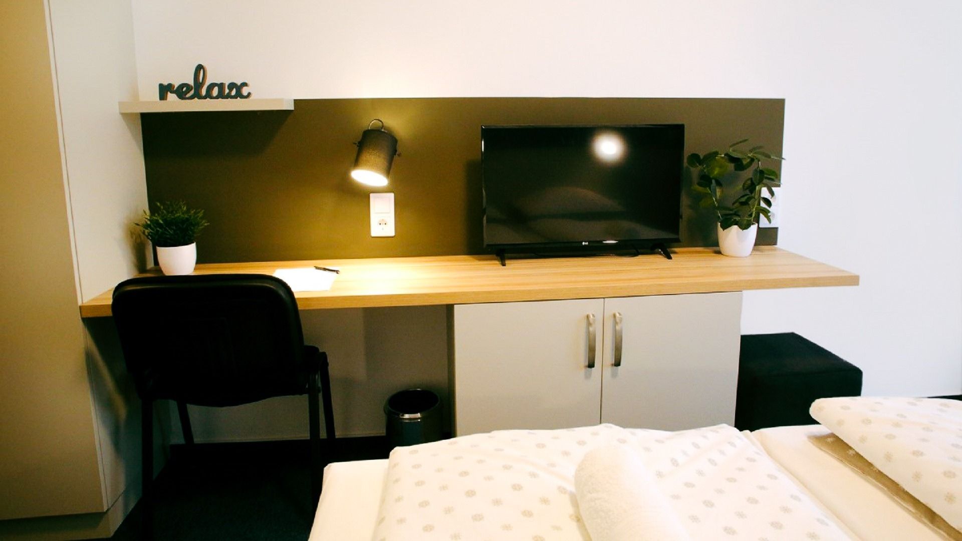 Schreibtisch mit Holzplatte, Fernseher, dekorativer Schriftzug „Relax“ und zwei kleine Pflanzen, ideal für Gäste, die arbeiten oder entspannen möchten.