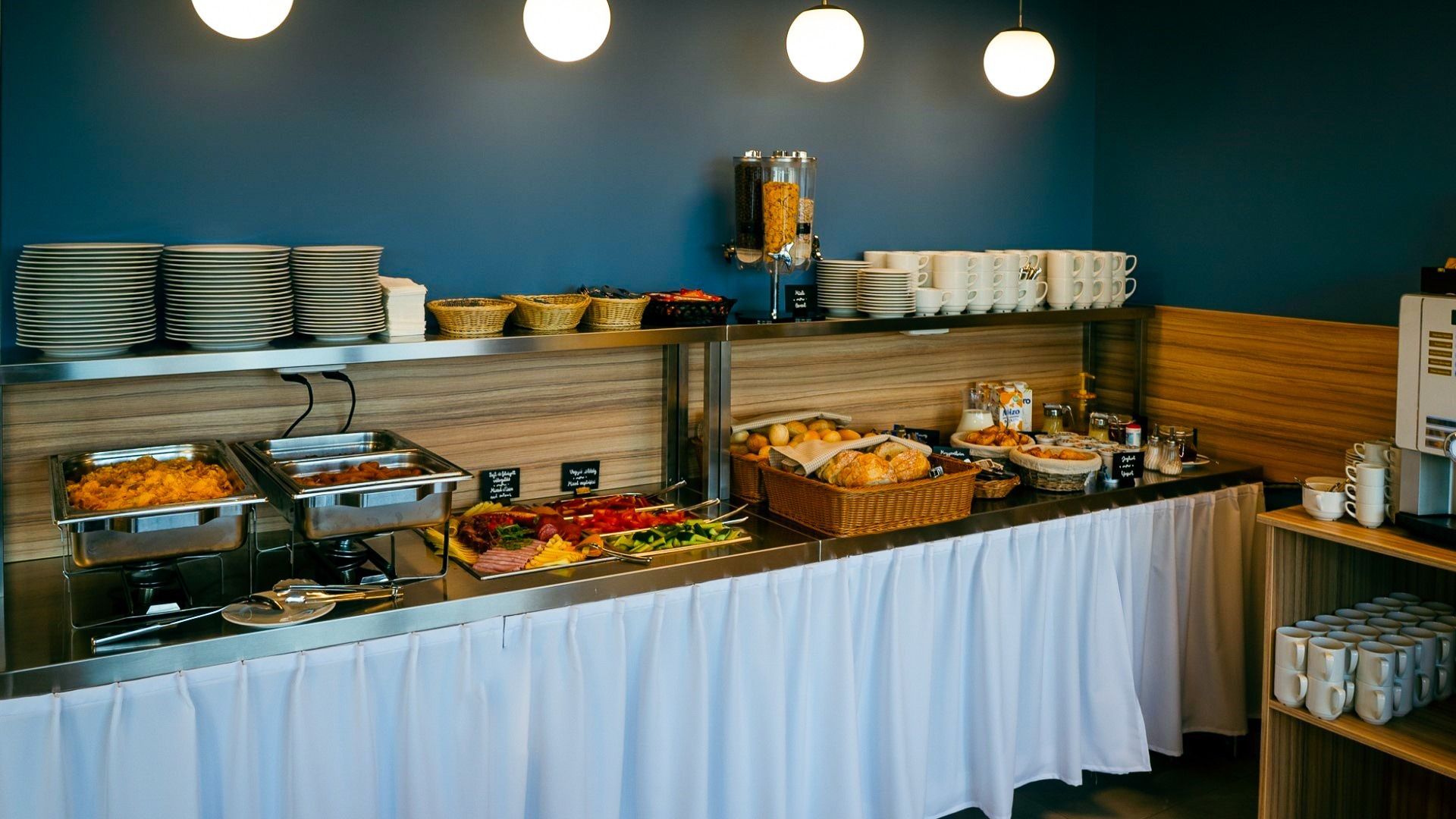 Ein reichhaltiges Frühstücksbuffet mit warmen und kalten Speisen, Brotkorb, Aufschnitt und frischen Früchten, präsentiert vor einer blauen Wand mit modernen Lampen.