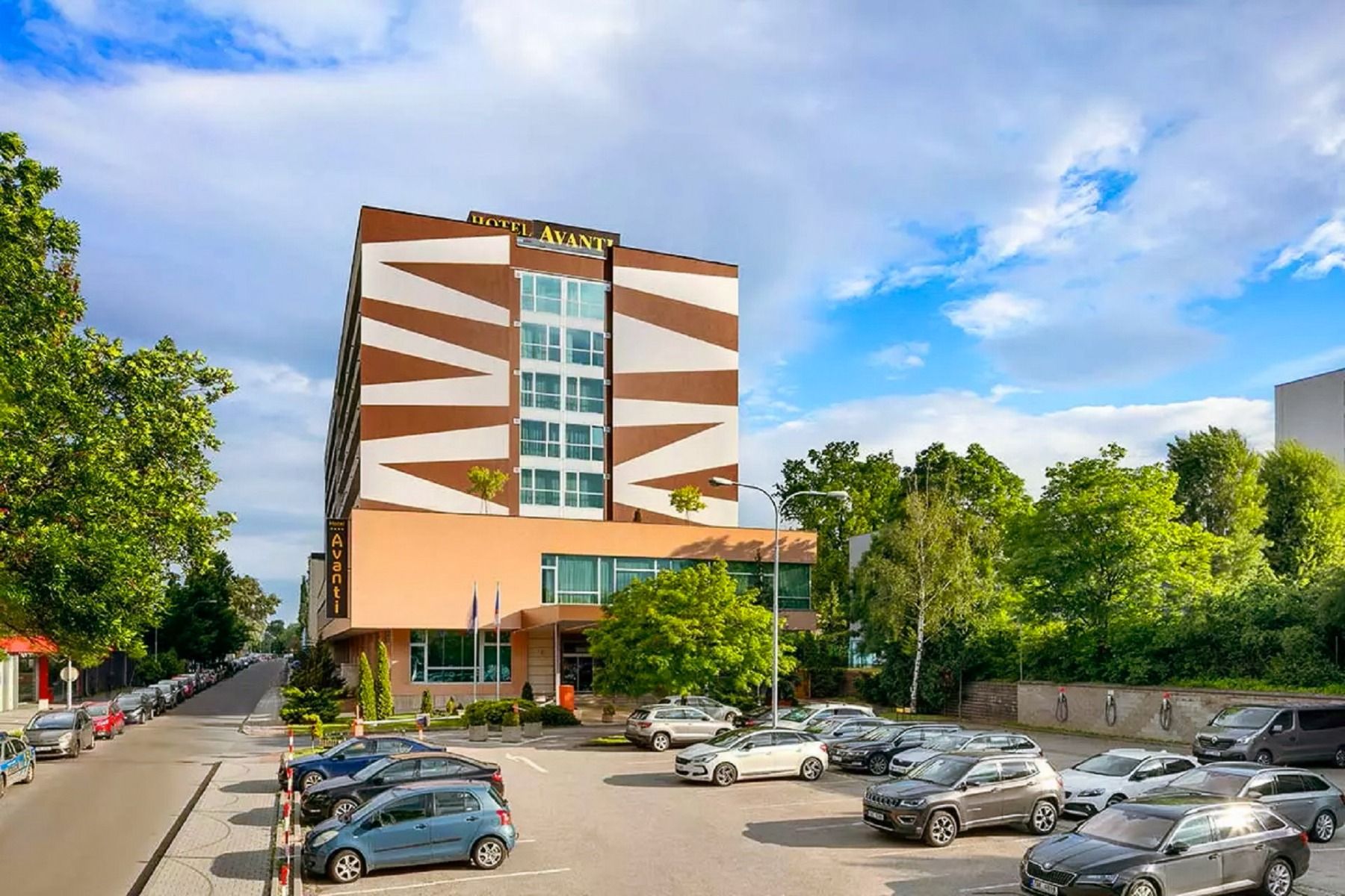 Ein modernes Hotelgebäude mit auffälligen geometrischen Mustern in Braun und Weiß, umgeben von grünen Bäumen und einem großen Parkplatz im Vordergrund.