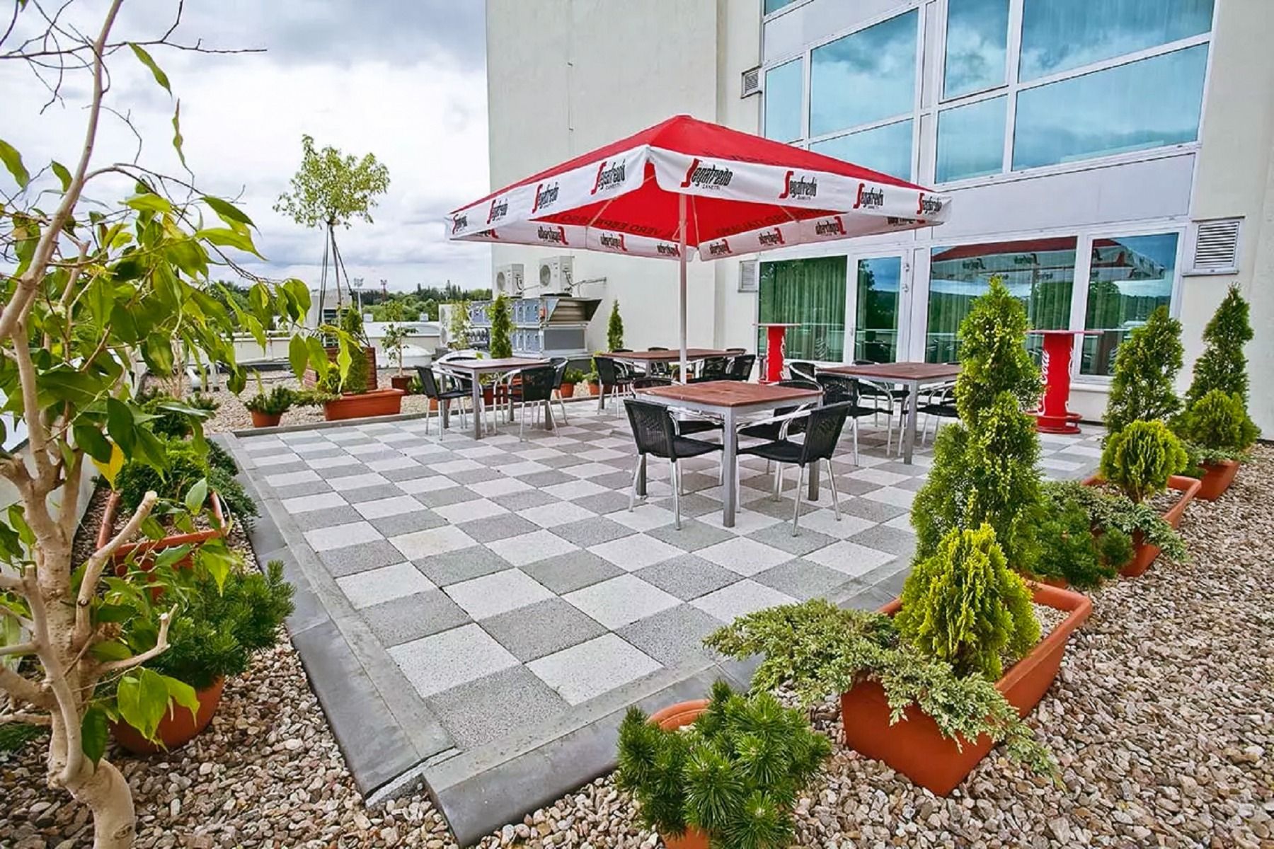 Eine gemütliche Terrasse mit gepflastertem Boden, modernen Sitzmöbeln und einem großen Sonnenschirm, umgeben von dekorativen Pflanzen und Bäumen.