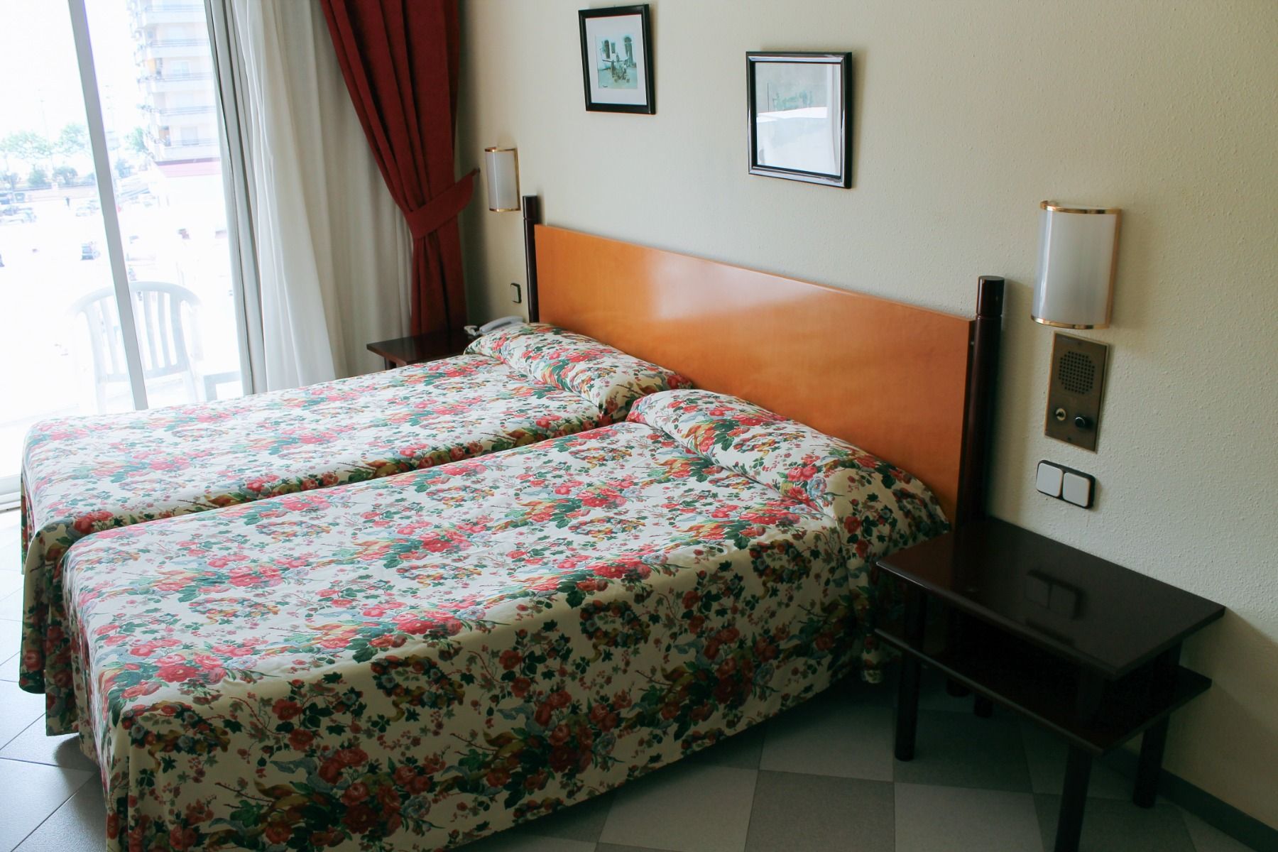 Hotelzimmer mit einem Doppelbett, floralem Bettüberwurf und großen Fenstern mit Vorhängen.