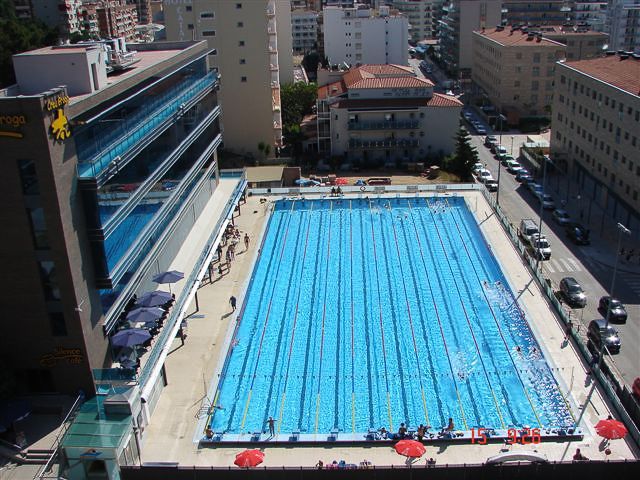 Vogelperspektive auf ein großes Freibad mit Schwimmbahnen, umgeben von städtischen Gebäuden und Sonnenschirmen.