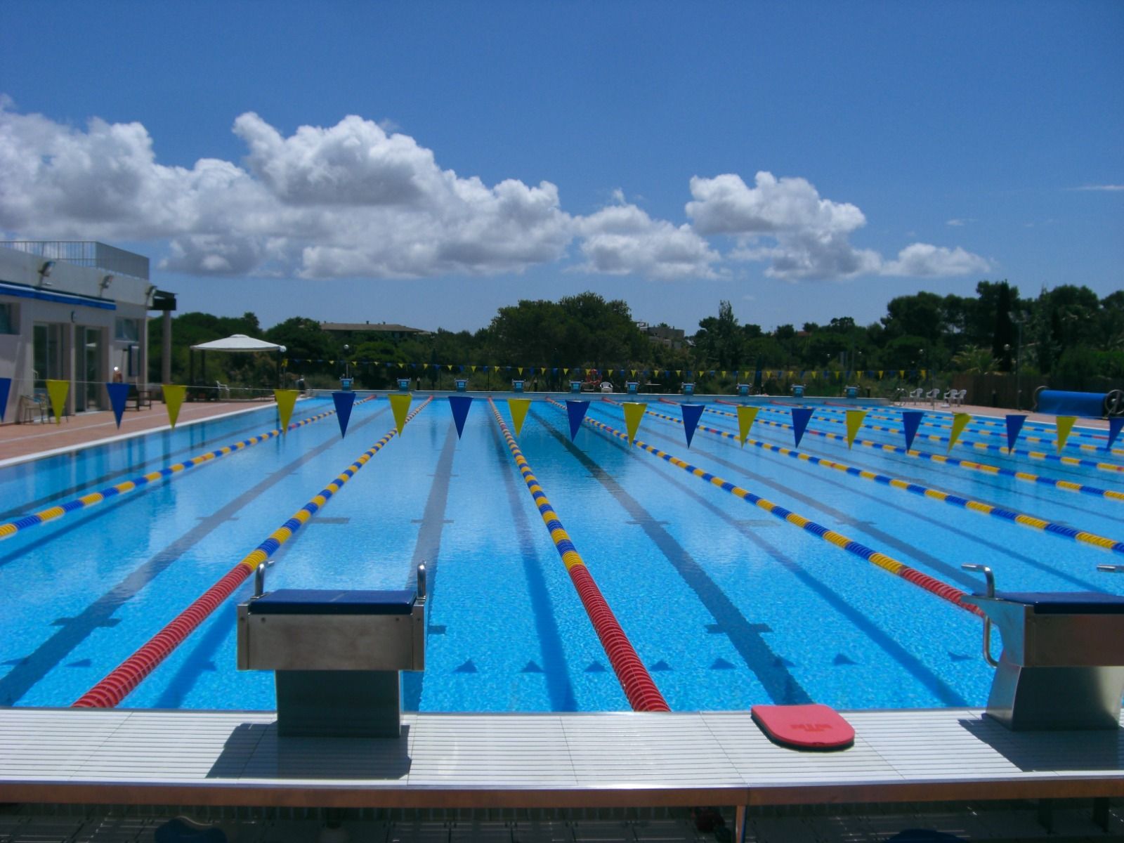 Außenansicht eines Schwimmbeckens mit abgetrennten Bahnen, Startblöcken und blauen und gelben Markierungsfahnen, unter freiem Himmel mit blauem Himmel und Wolken.