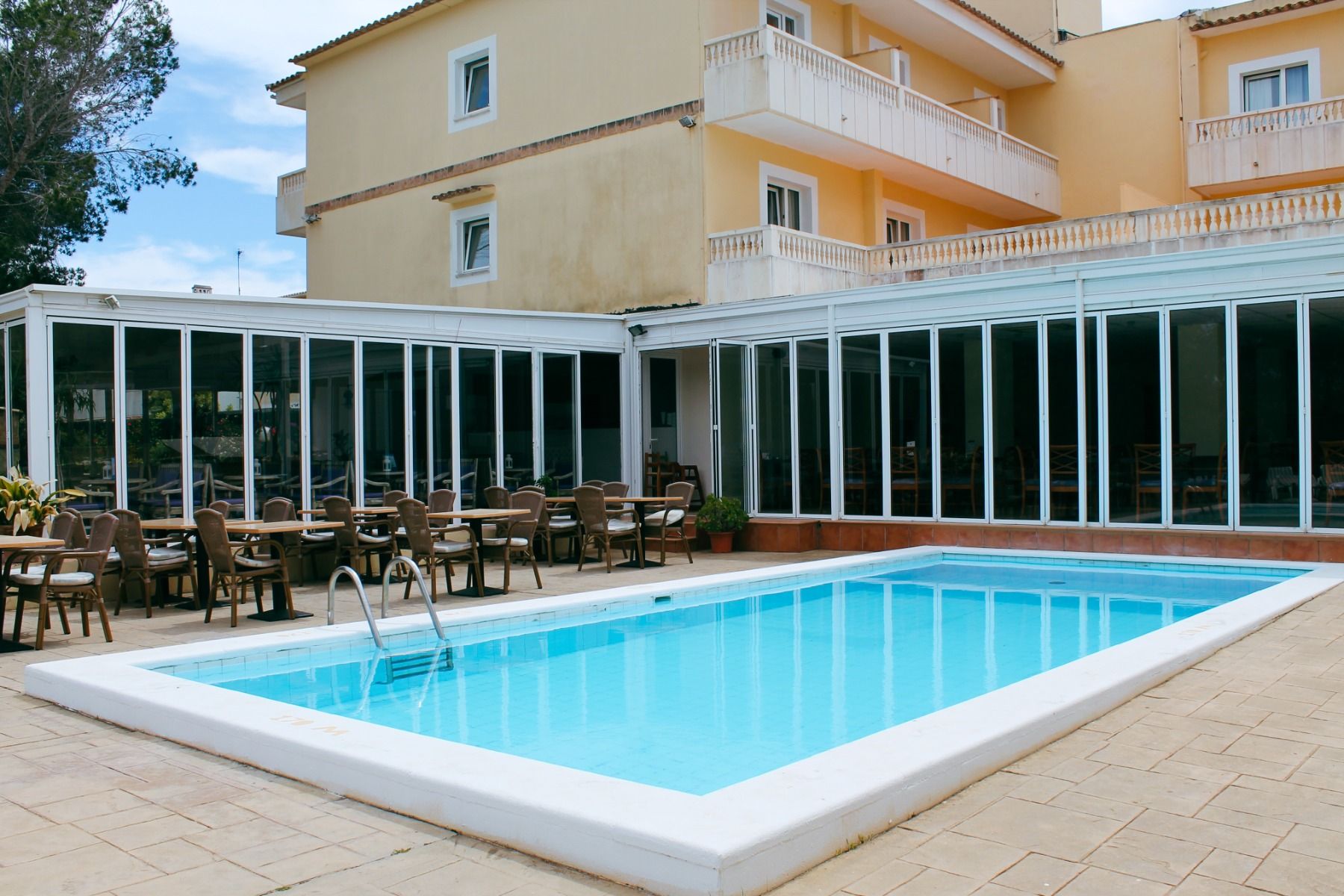 Hotelterrasse mit Pool, umgeben von Liegestühlen und Tischen mit Stühlen, angrenzend an ein gelbliches Gebäude mit Balkonen und großen Glasfenstern.