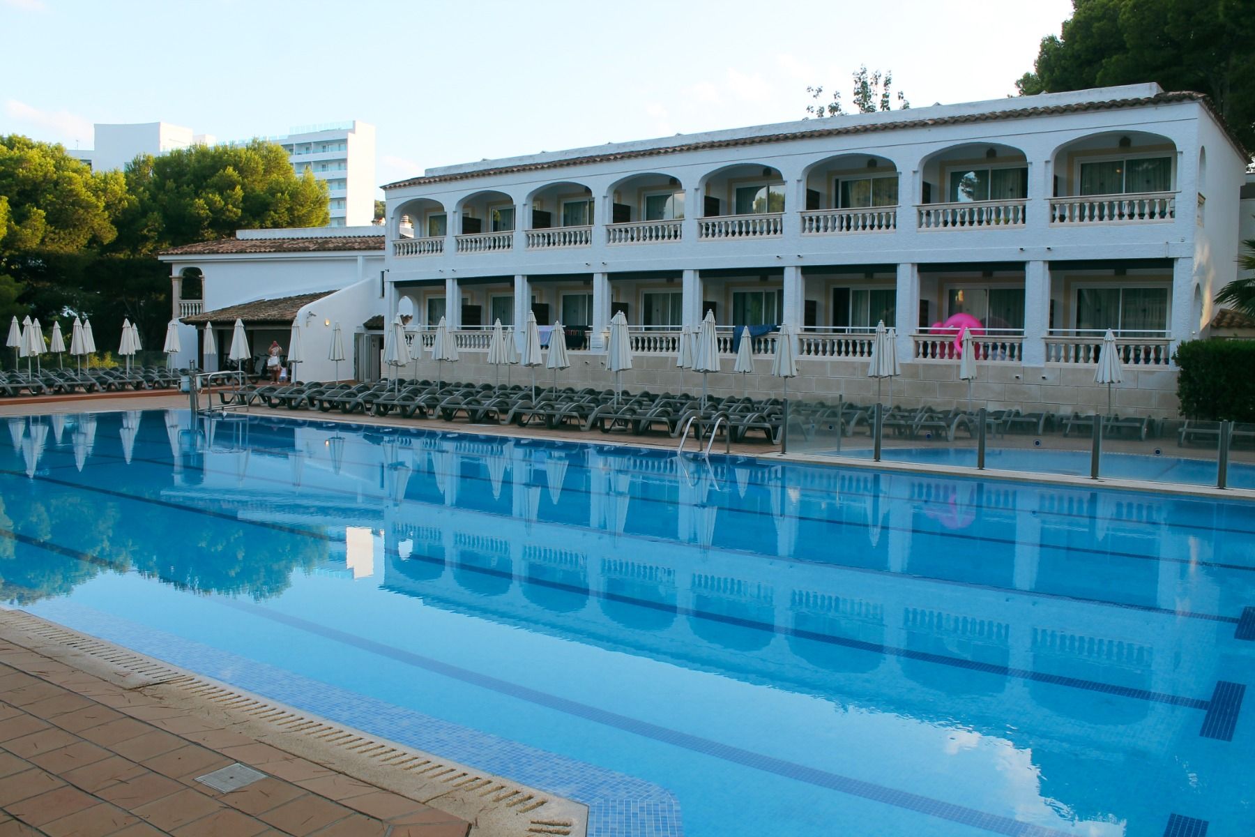 Großer Außenpool mit klar blauem Wasser, umgeben von weißen Hotelbalkonen und zahlreichen Sonnenliegen unter geschlossenen Sonnenschirmen.