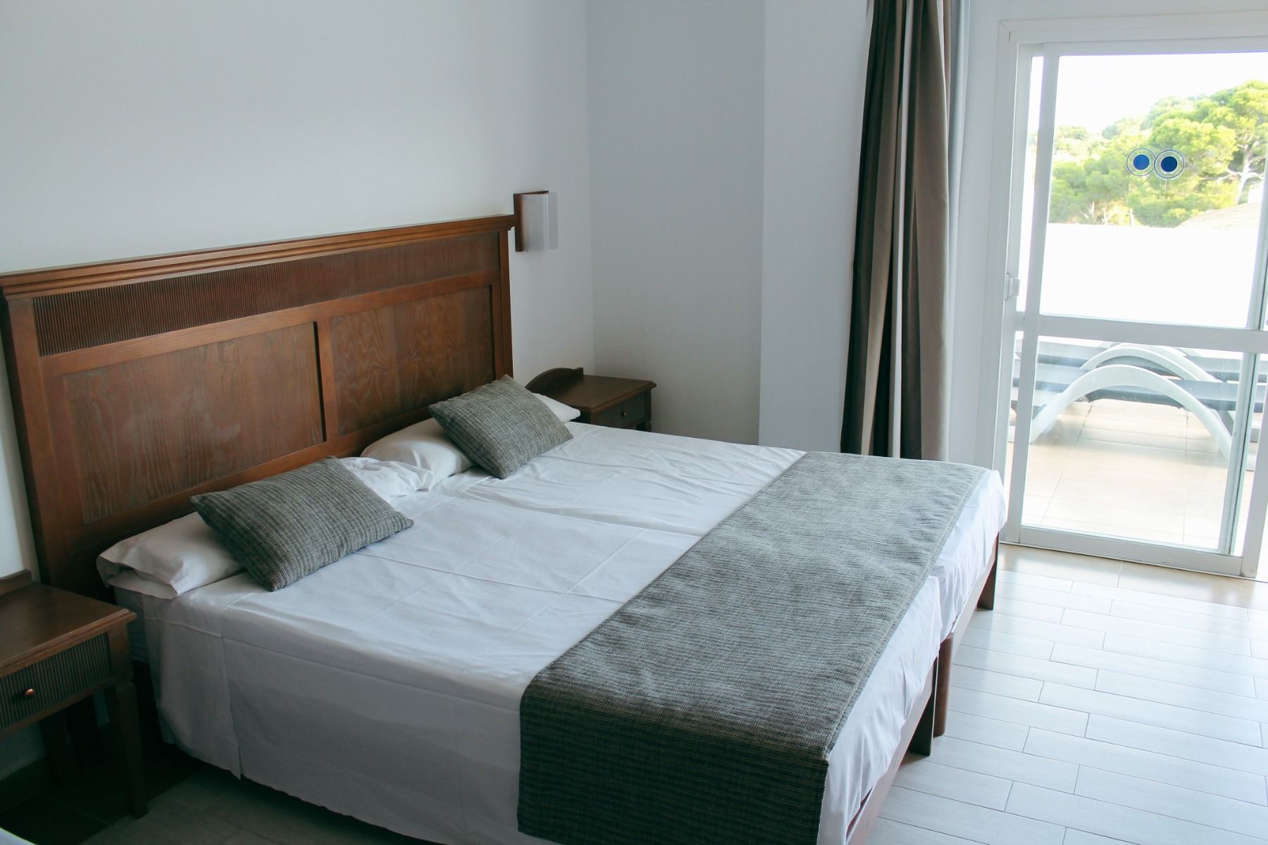 Helles Hotelzimmer mit einem Doppelbett, grauen Kissen und einer großen Fensterfront mit Zugang zu einem Balkon mit Liegestühlen.