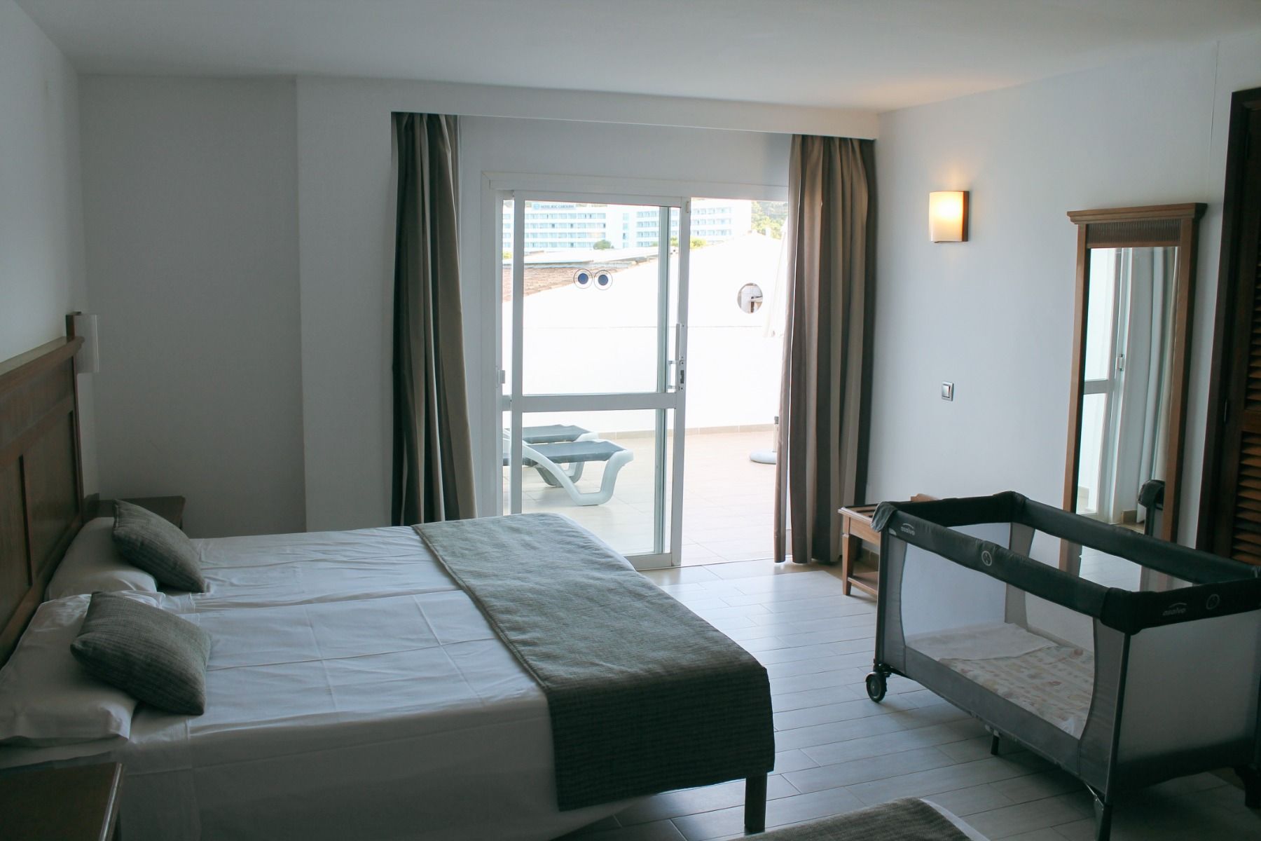 Modernes Hotelzimmer mit einem Doppelbett, einer Terrasse mit Sonnenliegen und einem Babybett in einer geräumigen, minimalistischen Einrichtung.