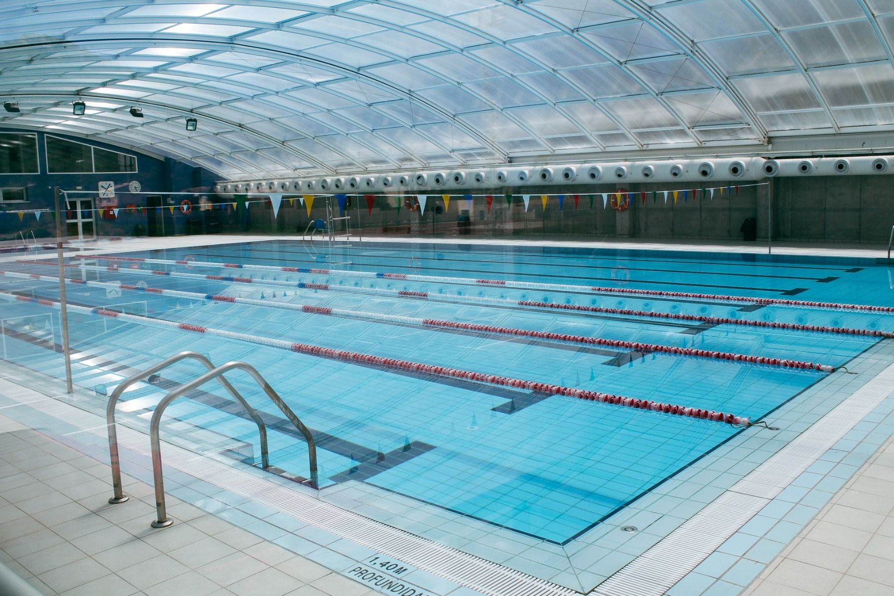 Überdachtes Schwimmbecken mit mehreren Bahnen, Edelstahl-Einstiegsleitern und einer Glasfassade, die für viel Tageslicht sorgt.
