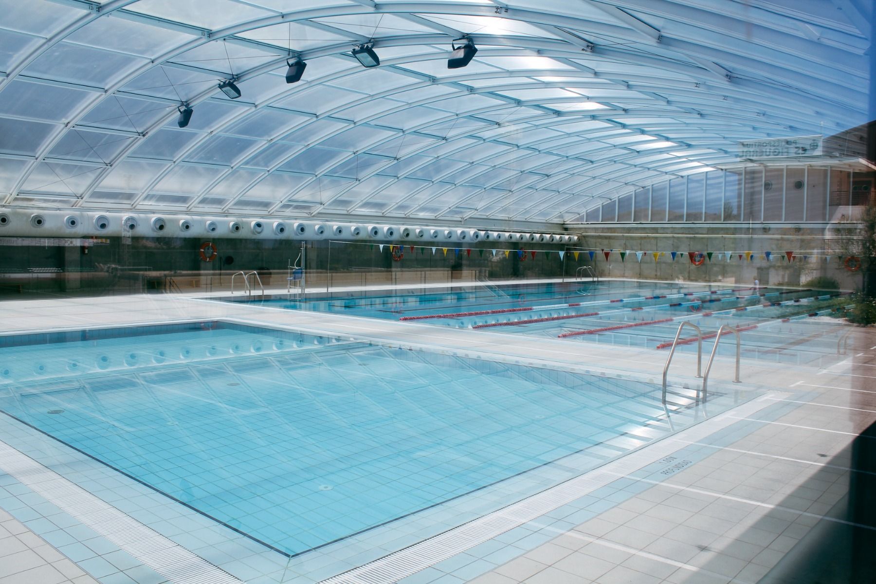 Großes, überdachtes Schwimmbecken mit mehreren Bahnen, umgeben von einer modernen Glaskuppel und bunten Wimpeln zur Dekoration.
