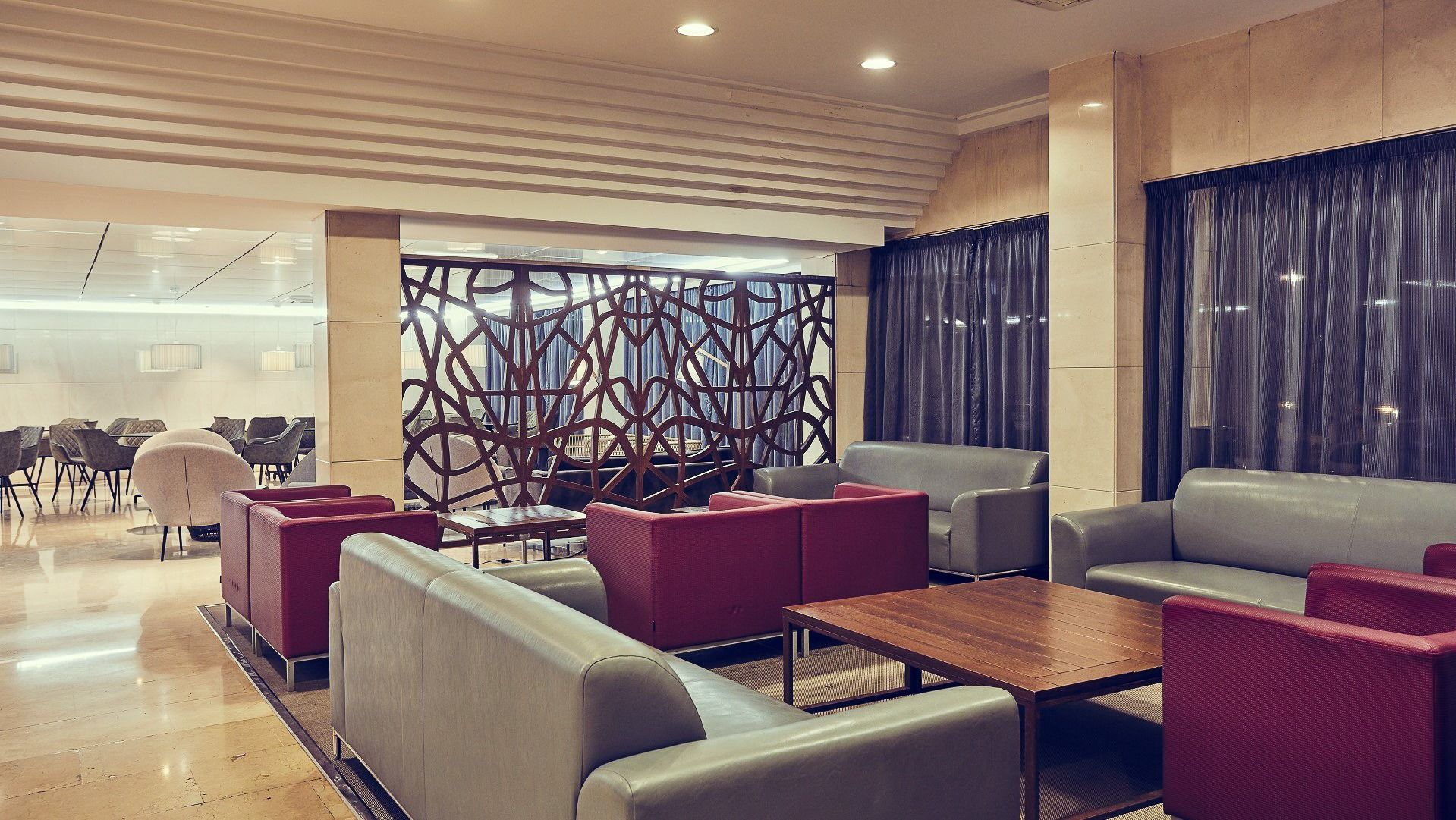 Hotel-Lounge mit roten und grauen Sesseln, Holztischen und moderner Trennwand.