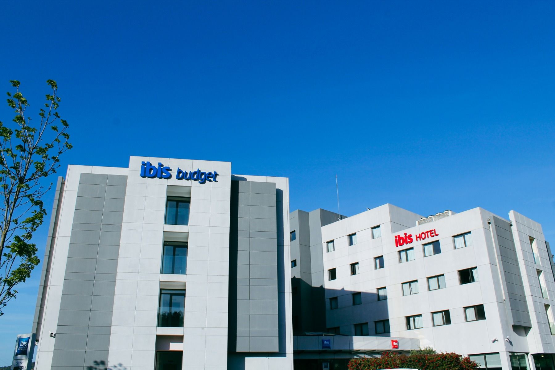 Außenansicht des Ibis Hotels mit moderner, grauer Fassade und blauer „Ibis Budget“- sowie roter „Ibis Hotel“-Beschriftung vor einem klaren blauen Himmel.