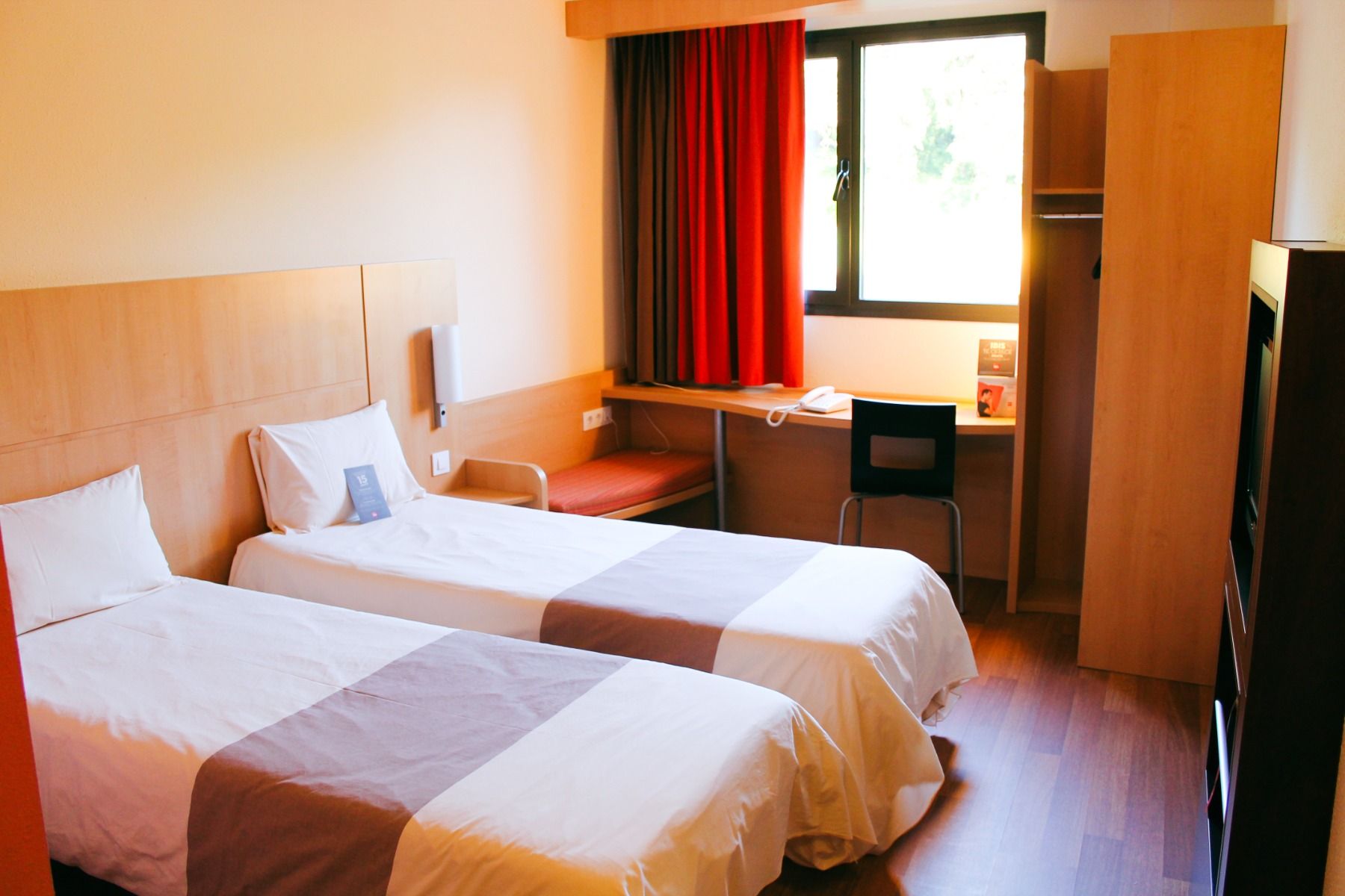 Hotelzimmer mit zwei Einzelbetten, weißen Bettdecken und einer schlichten Einrichtung mit Holzmöbeln und roten Vorhängen.