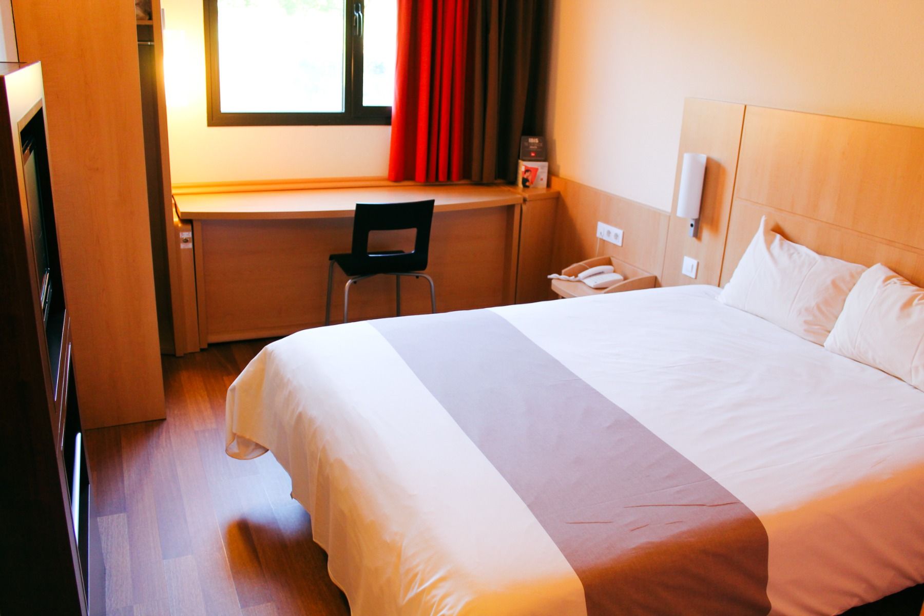 Hotelzimmer mit einem Doppelbett, hellbrauner Wandgestaltung, einem langen Schreibtisch und einem Fenster mit roten Vorhängen. 