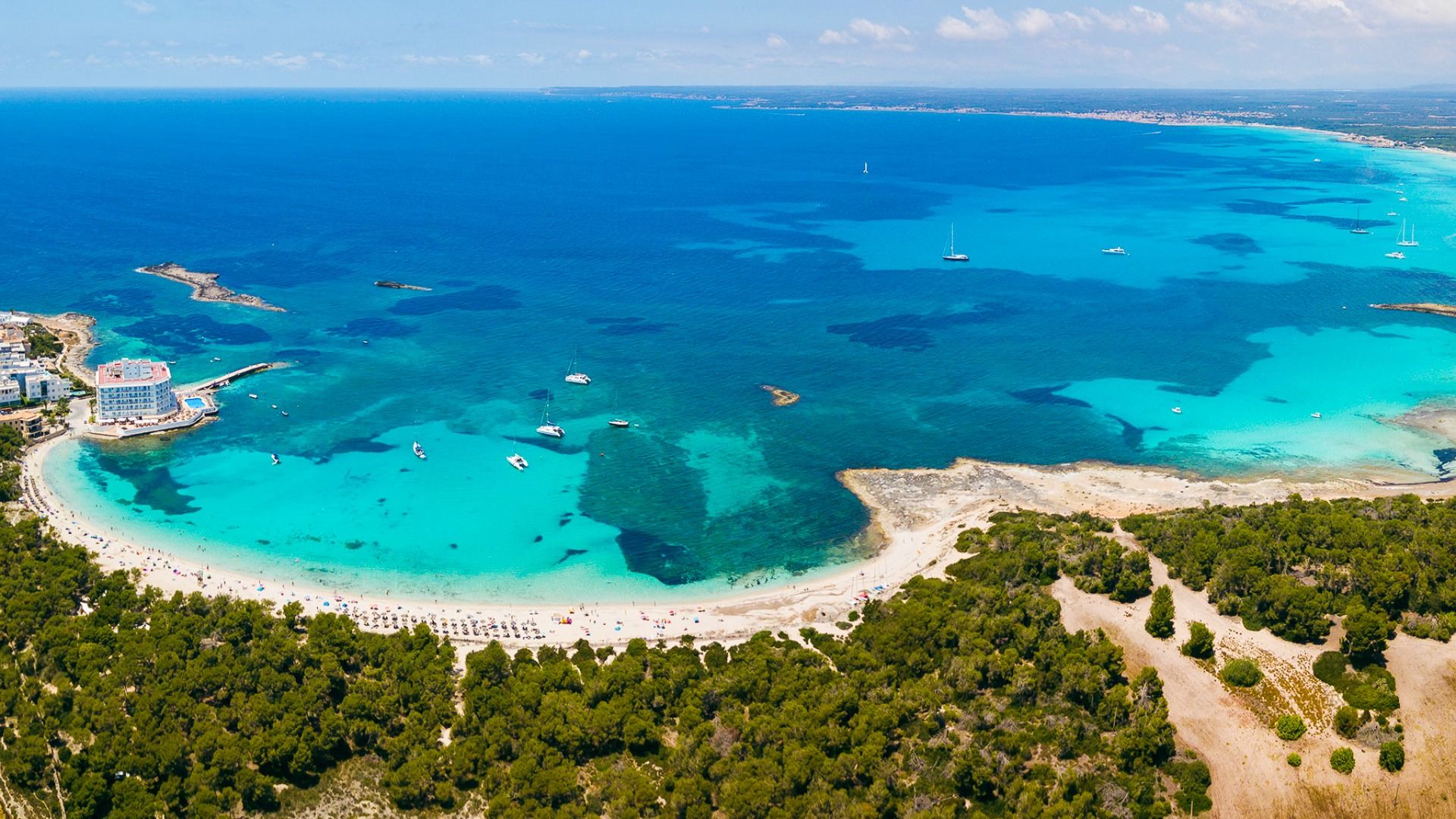 Luftaufnahme einer türkisblauen Bucht auf Mallorca mit Sandstrand, Segelbooten und umliegender grüner Landschaft