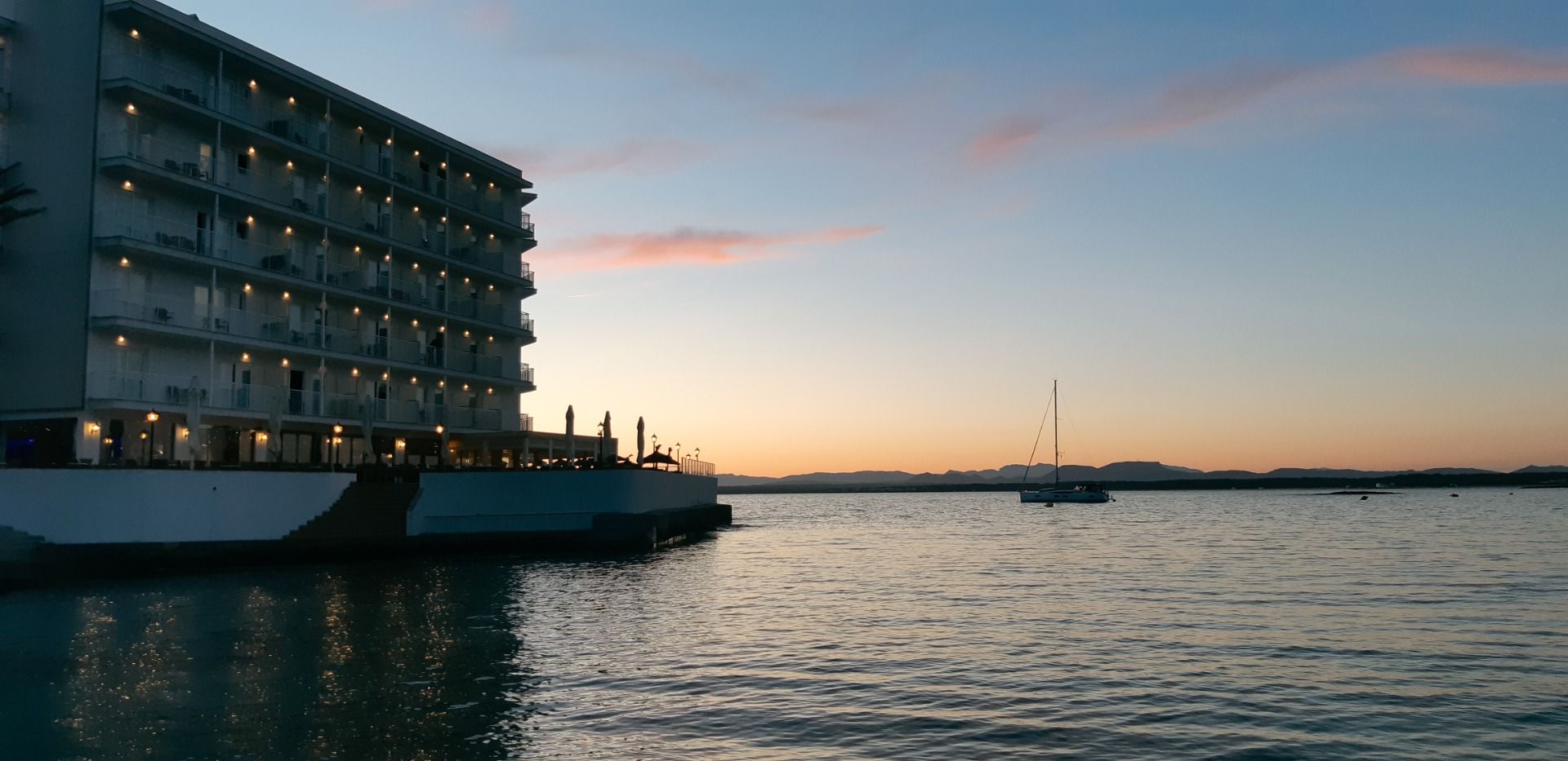 Hotel Marques bei Sonnenuntergang, mit beleuchteter Fassade direkt am Wasser und einem Segelboot im Hintergrund.
